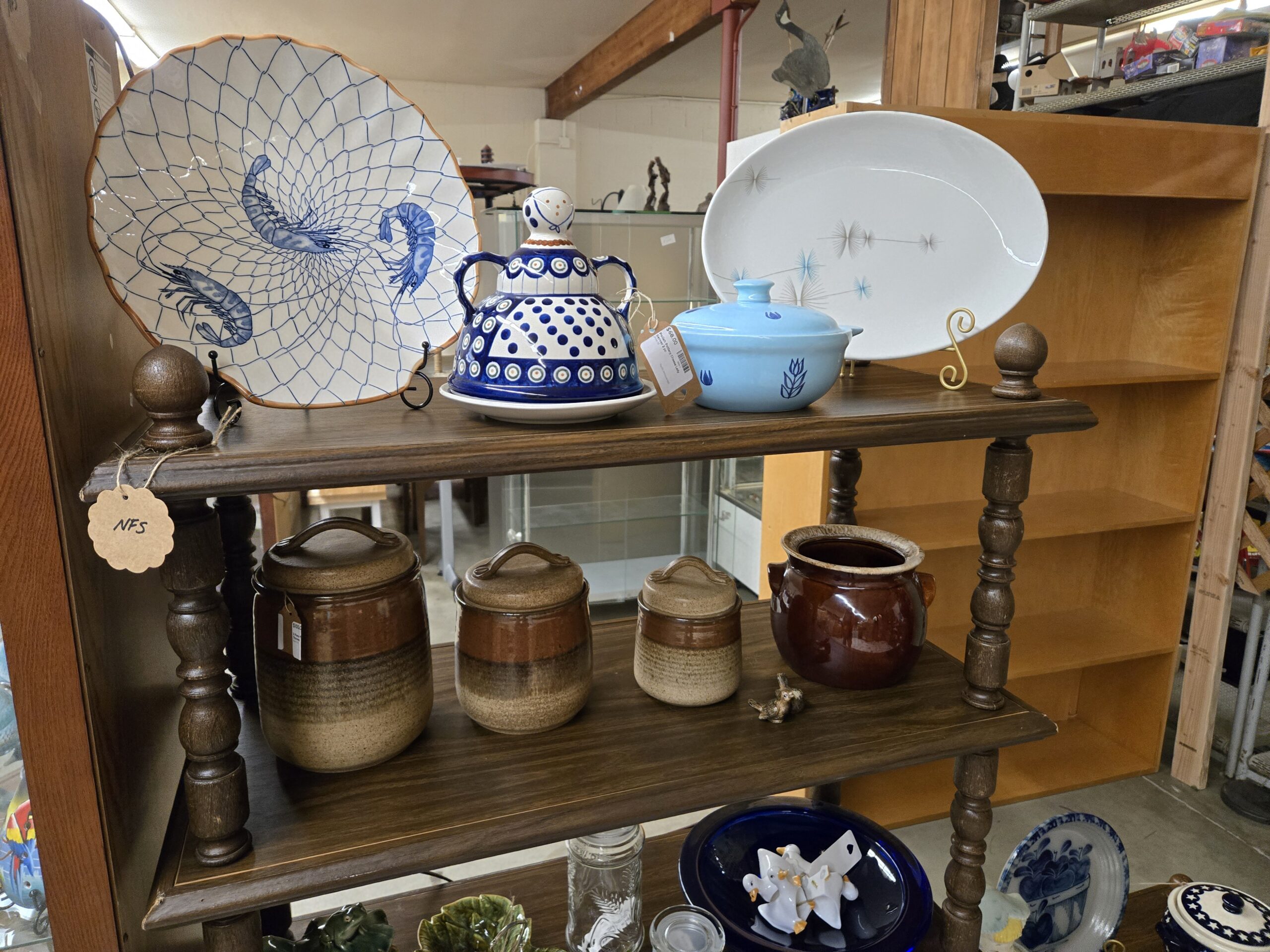 Scalloped blue and white plate painted with shrimp inside a fishing net pattern, a Polish pottery cheese dome in the Boleslawiec peacock-eye pattern, a light blue covered casserole with cobalt tulip decoration, a white oval platter with a mid-century starburst pattern, three lidded stoneware canisters in brown and tan bands, a brown drip-glaze bean pot, and a small brass bird figurine at Shop 55, Pacific Antiques Mall in Tacoma, WA.