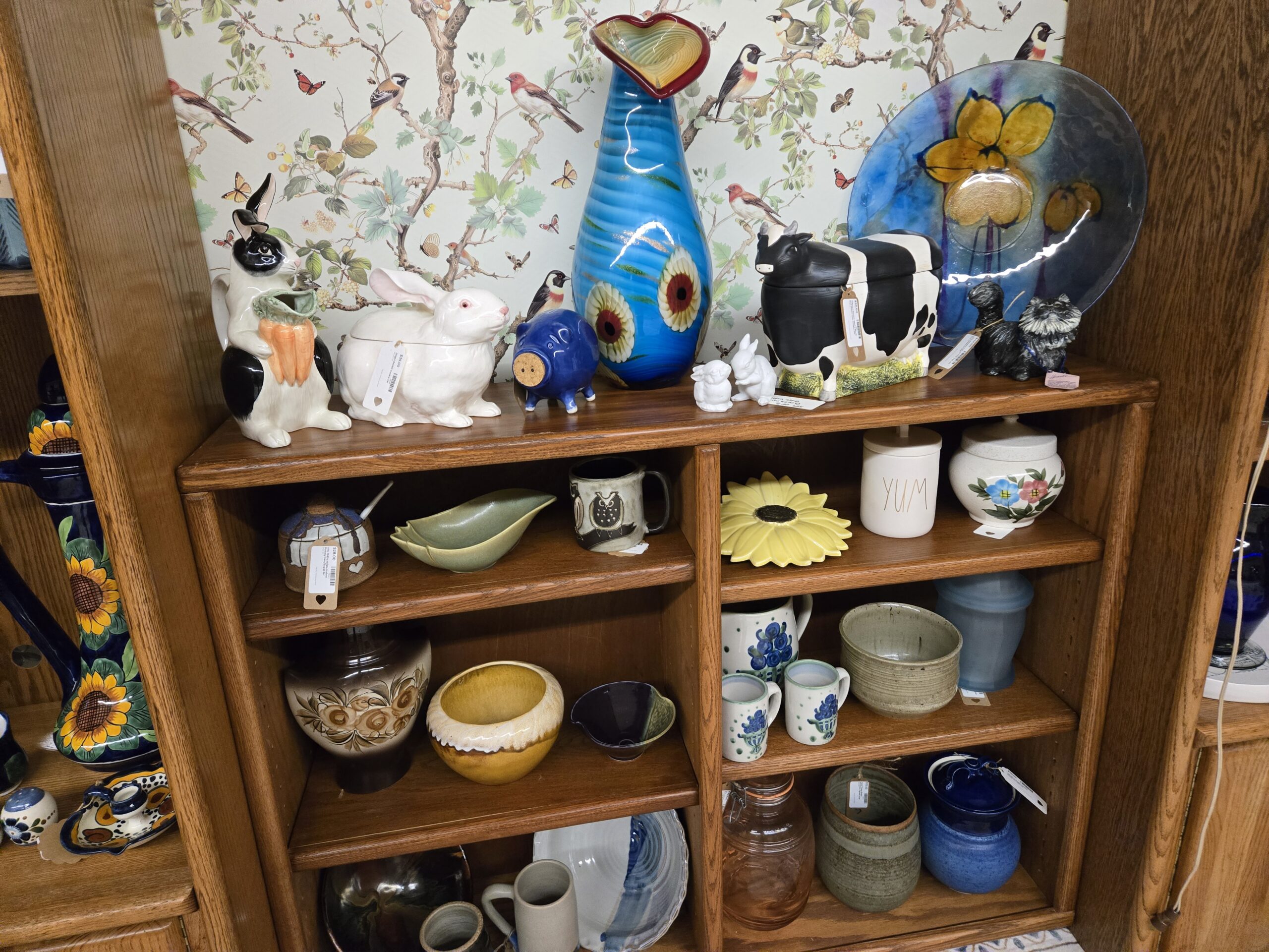 Black and white rabbit pitcher holding carrots, a white rabbit covered dish, a cobalt pig bank with cork stopper, a tall turquoise art glass vase with a heart-shaped red rim and floral cane decoration, small white bisque bunny figurines, a Holstein cow covered dish, a fused glass platter with yellow flowers on blue, black and gray terrier figurines, a stoneware honey pot with dipper, a sage green leaf dish, an owl mug, a yellow sunflower dish, a Rae Dunn YUM canister, a floral painted covered jar, drip-glaze vases and bowls, a stoneware pitcher and mugs painted with blue flowers, hand-thrown stoneware bowls and crocks, a pink glass canister with bail lid, and a cobalt glazed covered jar at Shop 55, Pacific Antiques Mall in Tacoma, WA.