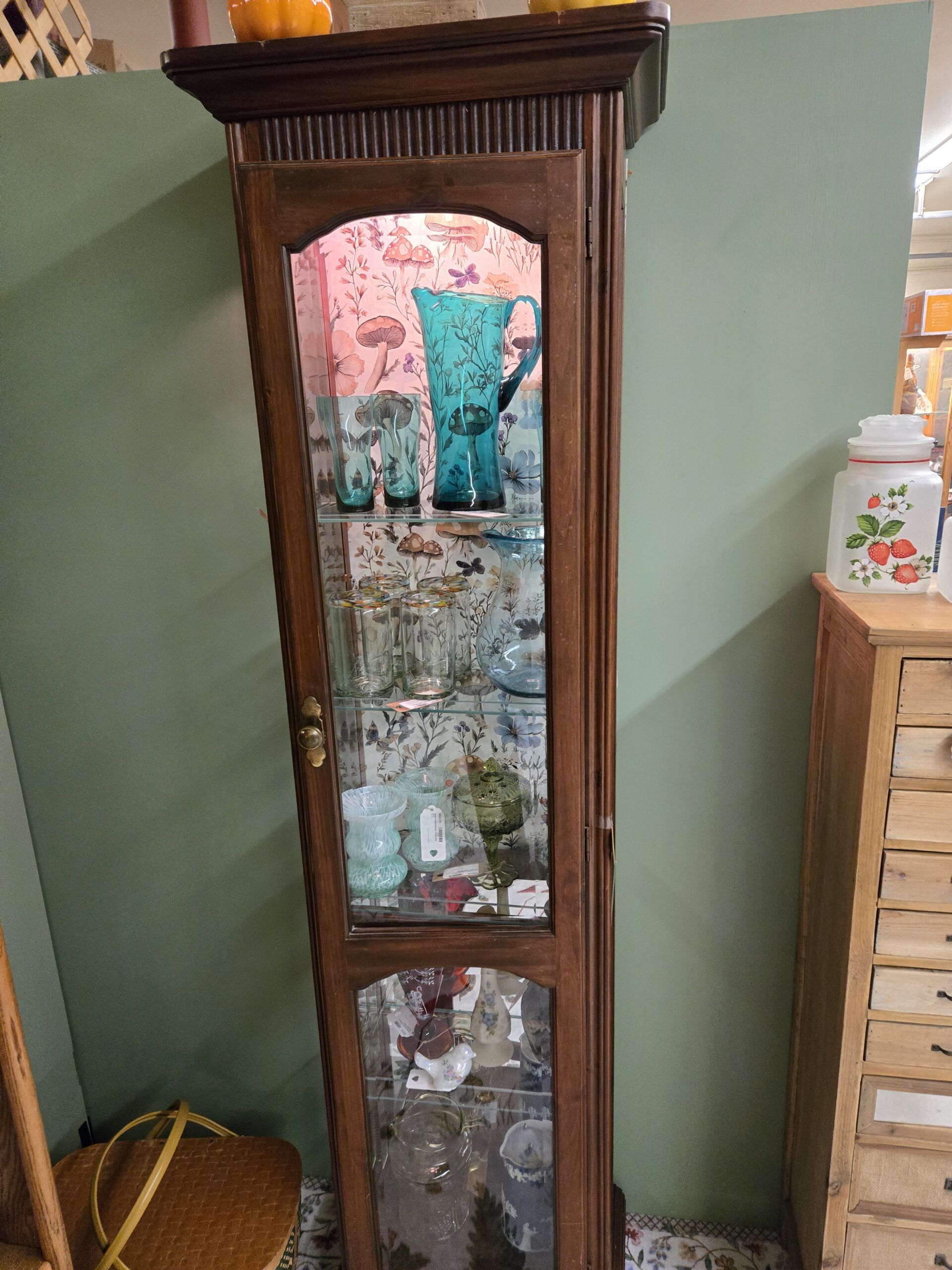 Lighted wood curio cabinet with mushroom and wildflower wallpaper backing holding a tall teal glass pitcher with matching tumblers, clear tumblers with multicolor confetti rims, a hand-blown pale blue pitcher, white and aqua spatter glass vases, an olive green footed glass covered dish, and on the lower shelves a white bird figurine, a floral painted bud vase, a blue and white relief pitcher, and clear glassware, beside frosted glass canisters painted with strawberries at Shop 55, Pacific Antiques Mall in Tacoma, WA.