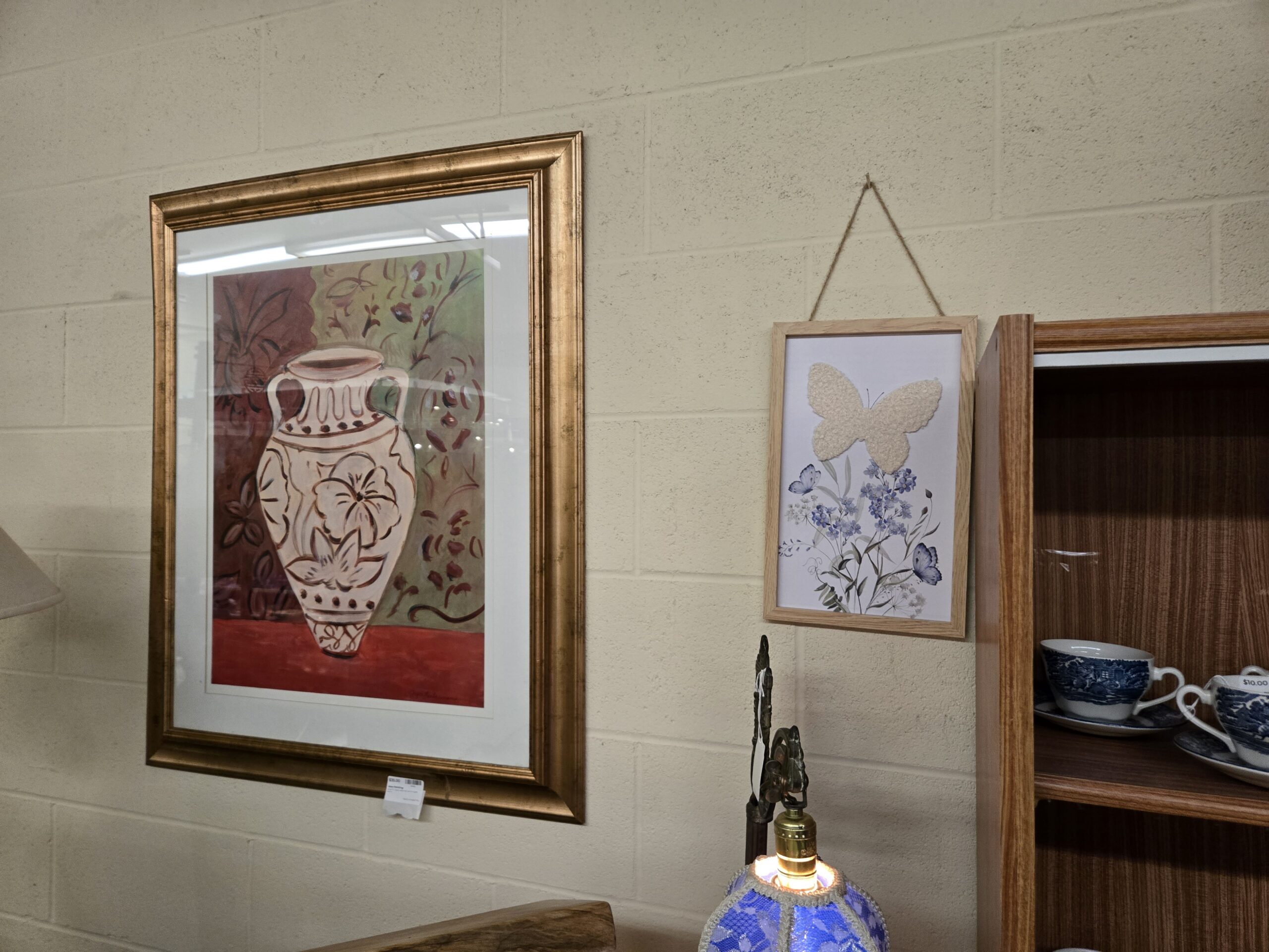 Matted print of a painted two-handled vase with floral motifs on a red and green ground in a gold frame, a wood-framed botanical print of a textured cream butterfly over blue wildflowers on a jute hanger, a blue lace lamp shade, and Liberty Blue teacups at Shop 73, Pacific Antiques Mall in Tacoma, WA.