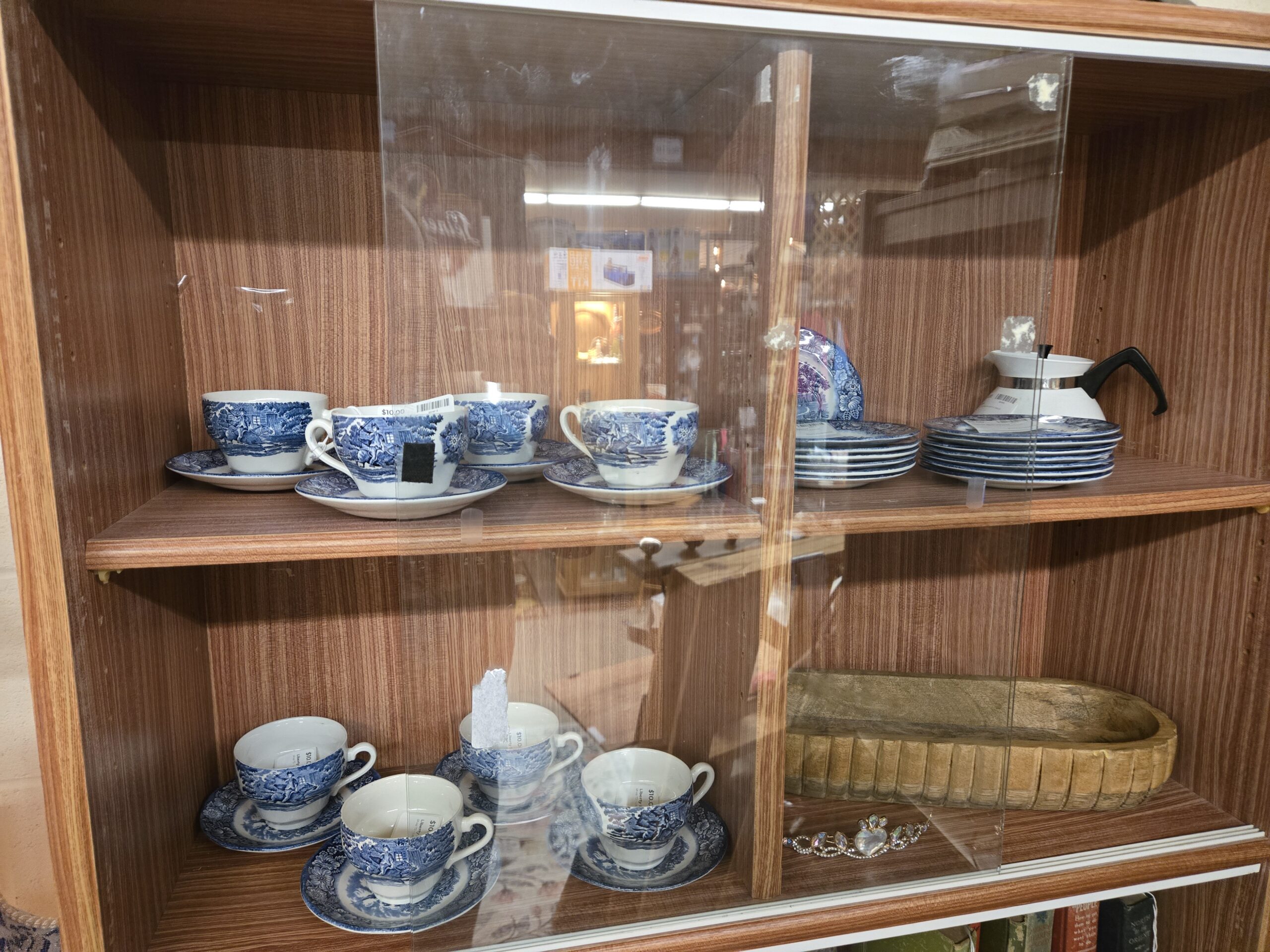 Liberty Blue transferware teacups and saucers showing Paul Revere's ride, stacks of matching blue and white plates, a transferware plate in blue and purple, a white stovetop coffee pot with a chrome band and black handle, a carved oblong wooden bowl, and a rhinestone tiara with a heart-shaped stone at Shop 73, Pacific Antiques Mall in Tacoma, WA.