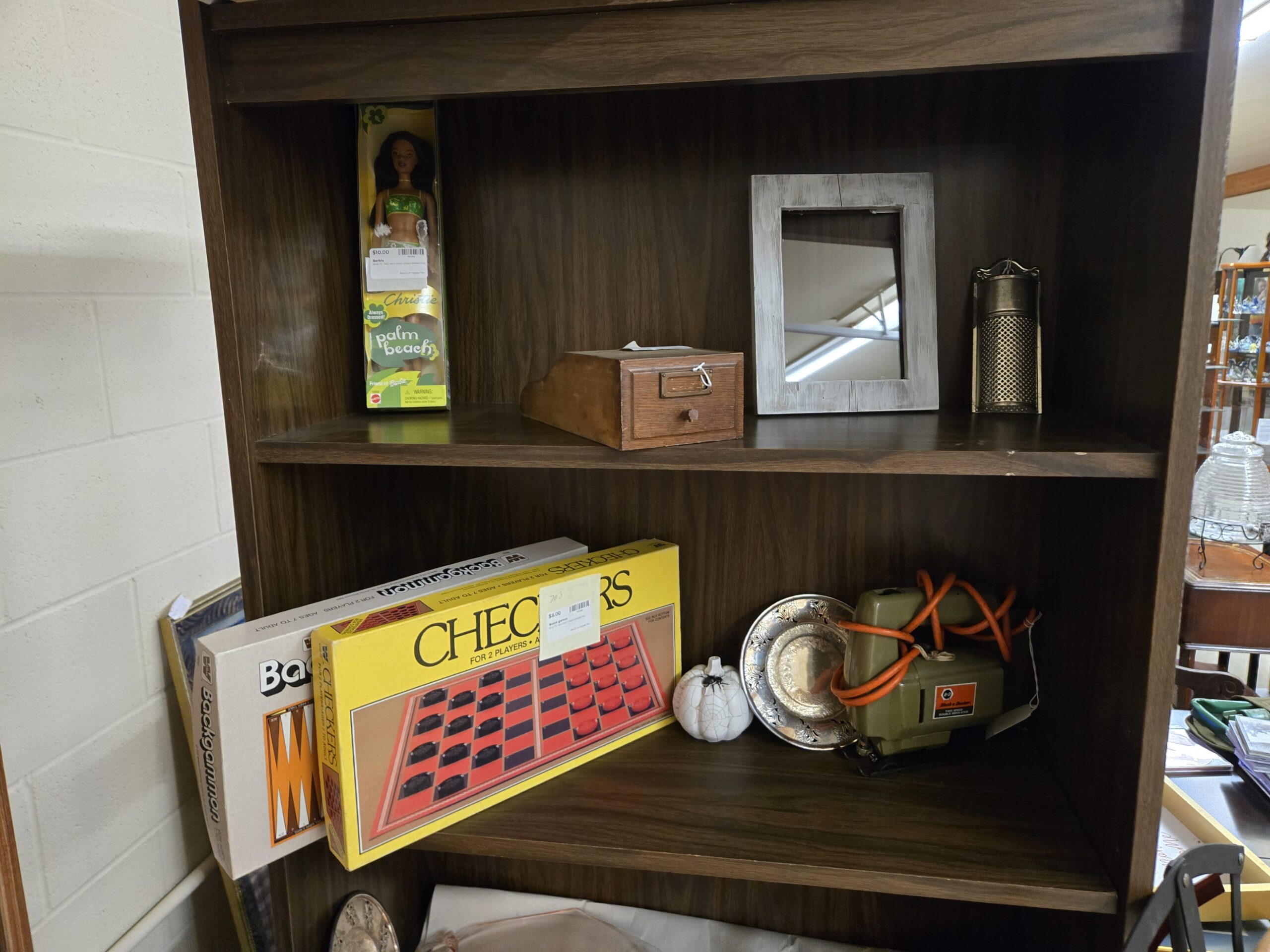 Boxed Mattel Palm Beach Christie doll, a wooden index card file box with a brass label holder and knob, a whitewashed wood-frame mirror, a hanging tin grater, boxed vintage Checkers and Backgammon games, a white ceramic pumpkin with spiderweb detail, a pierced silver-plate dish, and an olive green Black & Decker jigsaw with an orange cord at Shop 73, Pacific Antiques Mall in Tacoma, WA.