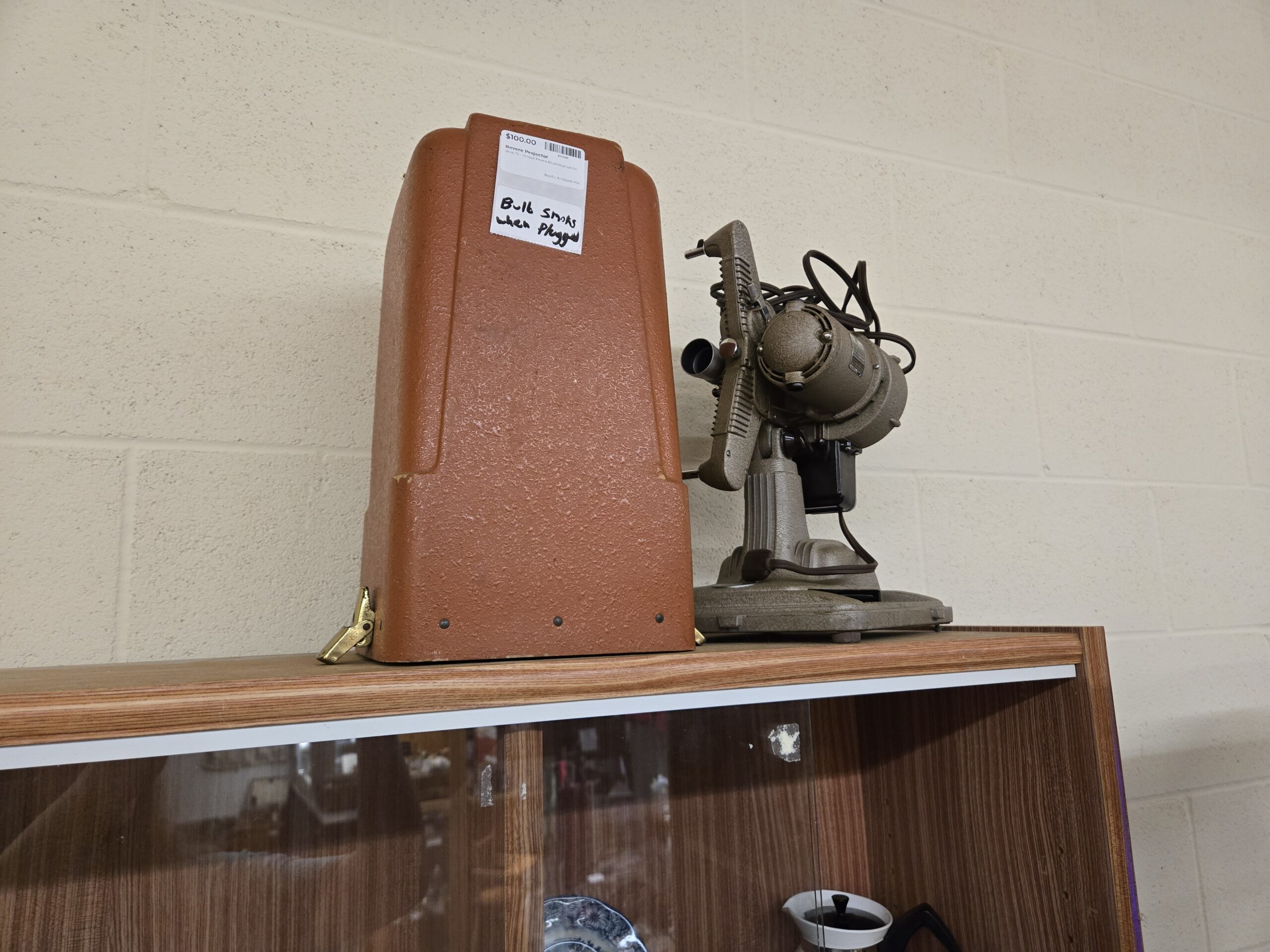 Vintage Revere movie projector in a gray-green crinkle finish with reel arms and a cloth-covered cord, beside its textured brown carrying case with a brass latch, at Shop 73, Pacific Antiques Mall in Tacoma, WA.