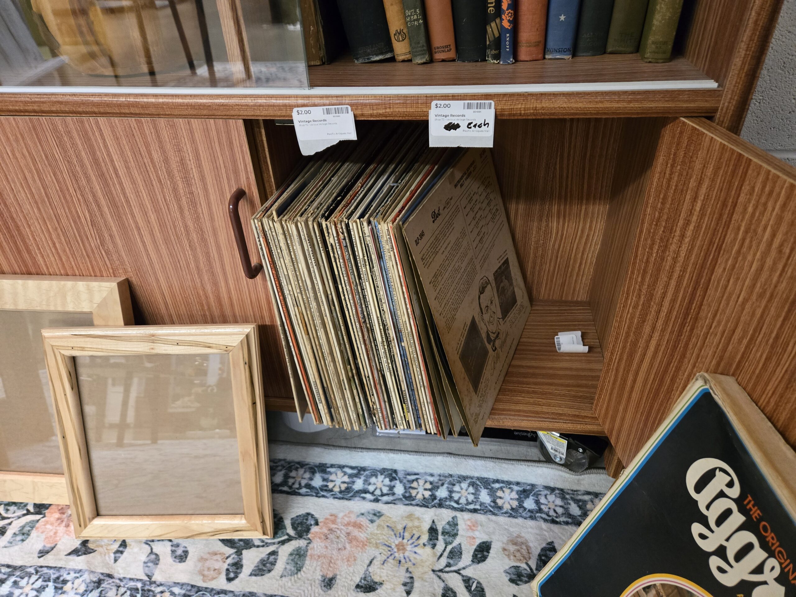Vintage vinyl LP records in a wood-grain cabinet, including a Billy Vaughn album on Dot Records, below a shelf of vintage hardcover books from Grosset & Dunlap, Knopf and Winston, with light wood picture frames leaning in front, at Shop 73, Pacific Antiques Mall in Tacoma, WA.