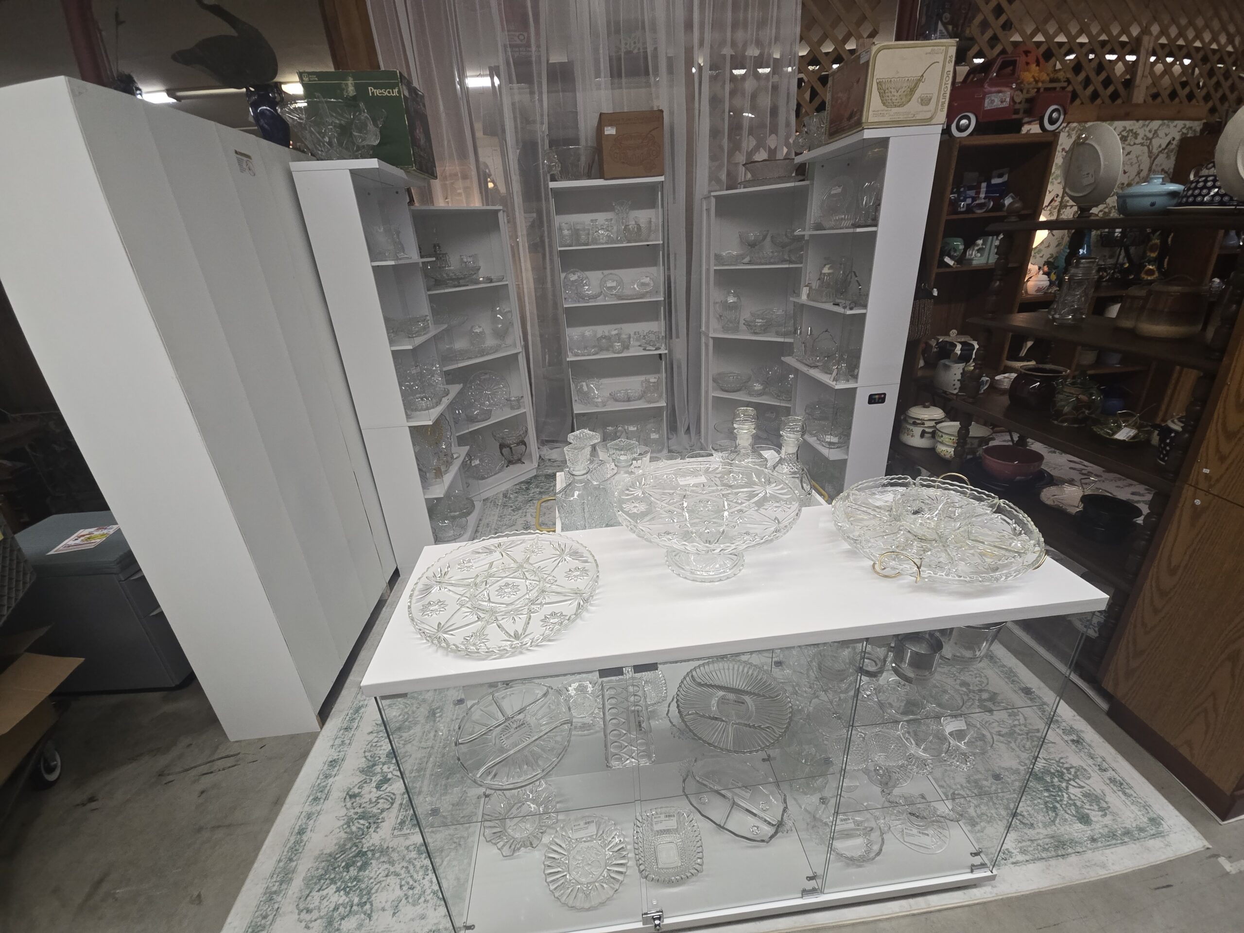 White glass-door shelving units filled with clear pressed glass bowls, compotes, pitchers and relish dishes, with boxed Prescut, Indiana Glass Princess and Anchor Hocking Arlington punch sets on top, plus a Star of David divided relish tray, a footed oval cake plate, a relish set in a brass-tone stand, and stoppered decanters at Shop 20, Pacific Antiques Mall in Tacoma, WA.