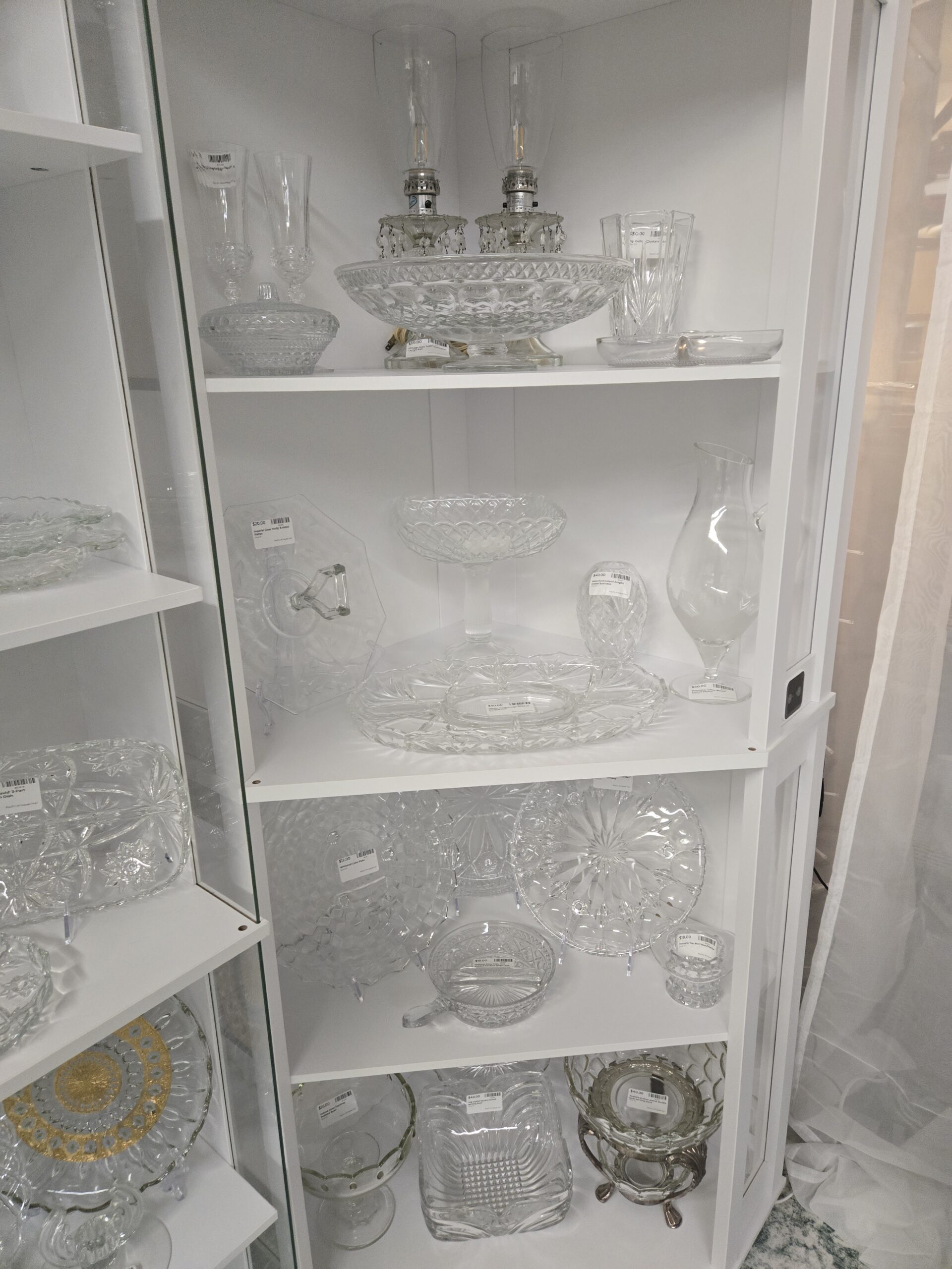Pair of prism-hung glass table lamps with hurricane chimneys, a footed diamond-pattern centerpiece bowl, champagne flutes, a covered candy dish, a cut crystal vase, an Imperial Glass Molly eight-sided center-handled platter, a lattice-rim pedestal compote, a Waterford Araglin crystal bud vase, a Rosenthal Lotus Blossom footed pitcher, a divided serving dish, a Whitehall cube-pattern cake plate, a Fostoria Top Hat vase, an Indiana Glass Teardrop pedestal bowl, a square slotted crystal serving bowl, and a two-bowl serving piece on a silver-plated stand at Shop 20, Pacific Antiques Mall in Tacoma, WA.