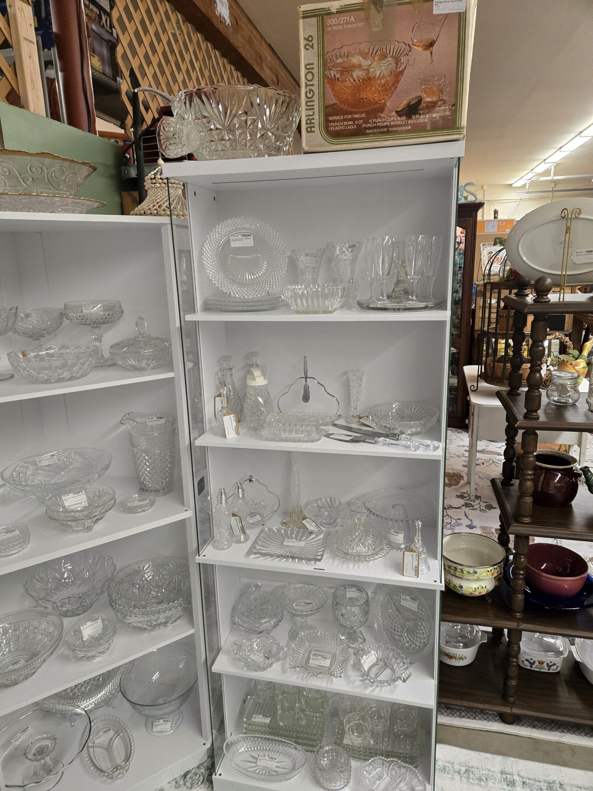 Anchor Hocking Arlington 26-piece punch set box beside a pressed glass punch bowl with cups, Miss America diamond point plates in a set of four, pressed glass vases, a ribbed oval bowl, champagne flutes on a glass tray, stoppered cruets, a Prescut syrup dispenser, a covered butter dish, a chrome-handled tidbit server, a bud vase, a star-pattern bowl, a crystal-handled cake knife and server, salt and pepper shakers with chrome tops, a chrome-handled basket, a swirl sugar and creamer, a beaded-edge cake stand, a covered butter dome, glass dinner bells, footed compotes, and stacked pressed glass tumblers at Shop 20, Pacific Antiques Mall in Tacoma, WA.
