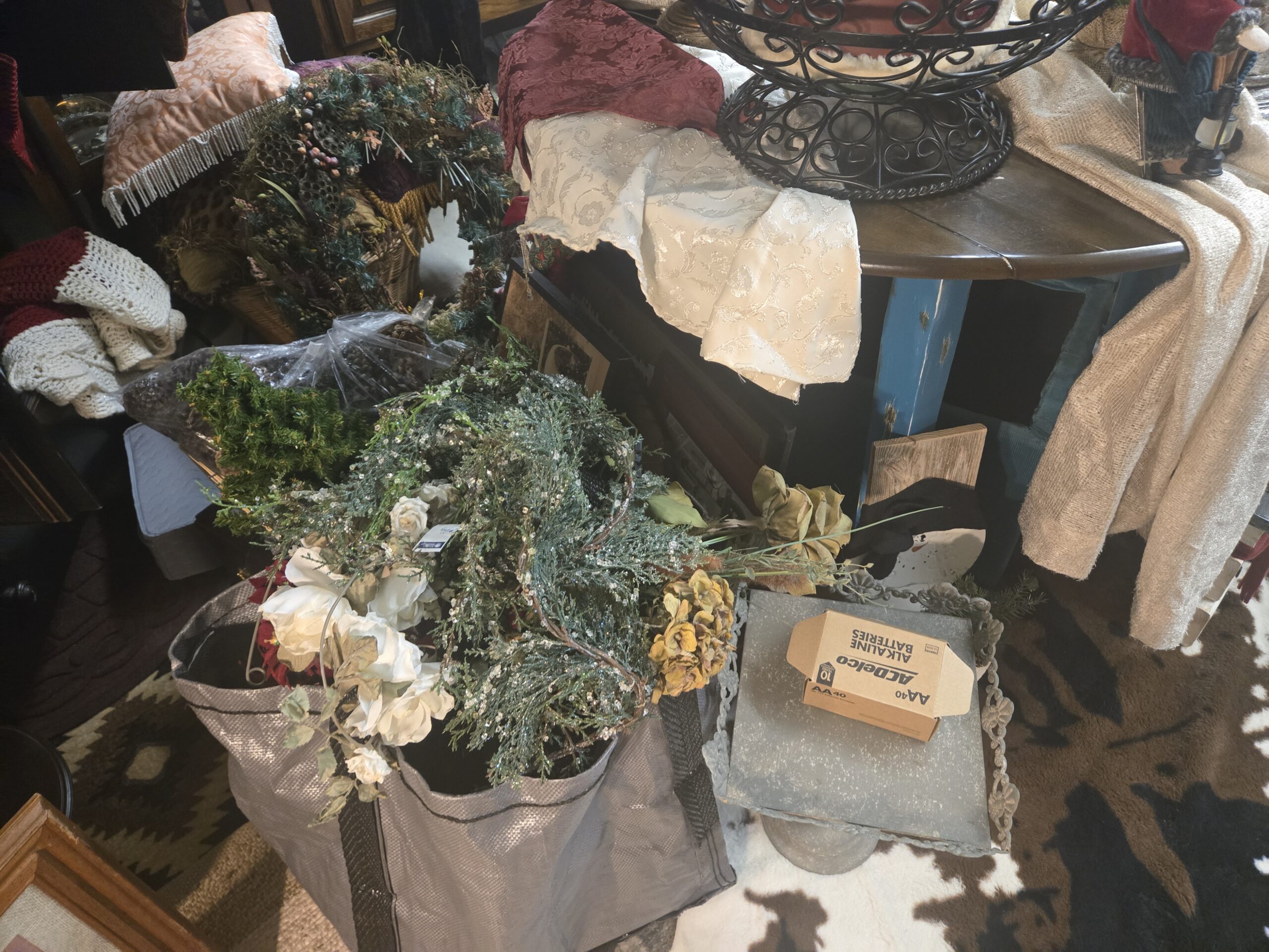 Glittered faux evergreen sprays and cream silk roses in a woven tote, gold dried-look hydrangea stems, a galvanized metal tray stand with scrolled edges, cream and silver damask fabric, burgundy damask fabric, a drop-leaf table with distressed blue legs, a black scroll wire centerpiece, faux wreaths, a peach fringed pillow, a red and cream crocheted throw, a painted snowman board, and a cowhide rug at Shop 217, Pacific Antiques Mall in Tacoma, WA.