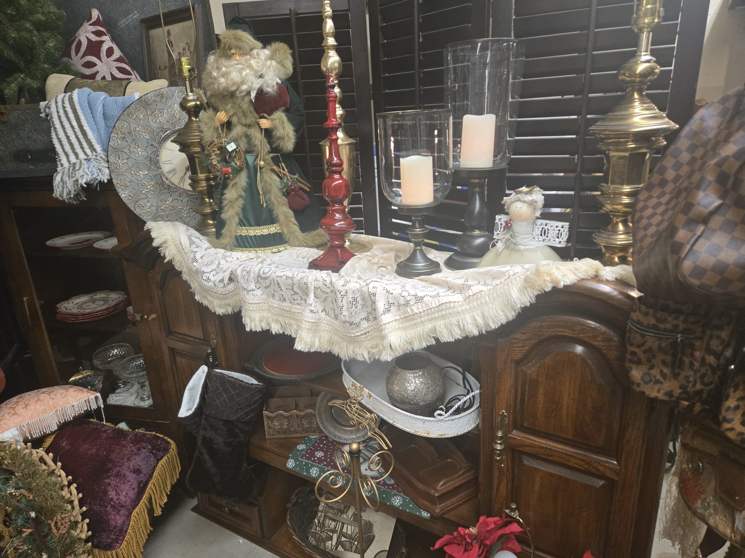 Father Christmas figure in a green robe with faux fur trim and a twig wreath, a red lacquered turned candlestick, brass table lamp bases, glass hurricane candle holders with LED candles, an angel figurine with silver wire wings, a fringed cream lace tablecloth, an embossed silver and gold fleur-de-lis wall clock with Roman numerals, a striped blue chenille throw, an oak buffet, a quilted brown stocking, a carved wood letter box, a gold wire reindeer, a white tray with a silver mercury glass sphere, and peach and purple fringed pillows at Shop 217, Pacific Antiques Mall in Tacoma, WA.