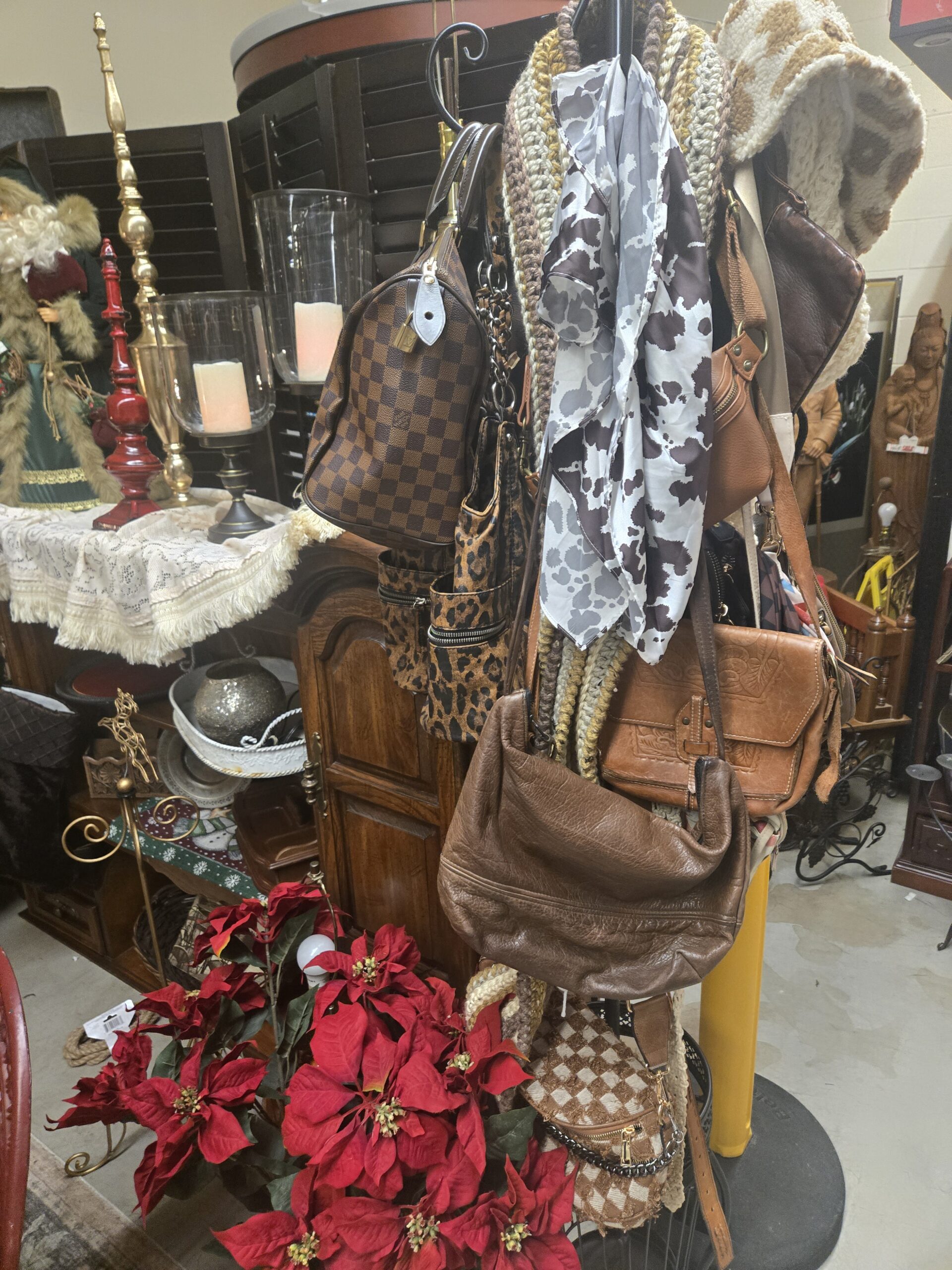 Handbag rack holding a brown checkerboard canvas satchel with a brass padlock, leopard-print bags, a cow-print scarf, crocheted scarves in brown, cream and gold, a tooled leather messenger bag, a brown leather hobo bag, and a checkered fabric belt bag with a chain strap, with red faux poinsettias, a Santa in a green robe, a red turned candlestick, a brass lamp, and glass hurricane candle holders at Shop 217, Pacific Antiques Mall in Tacoma, WA.