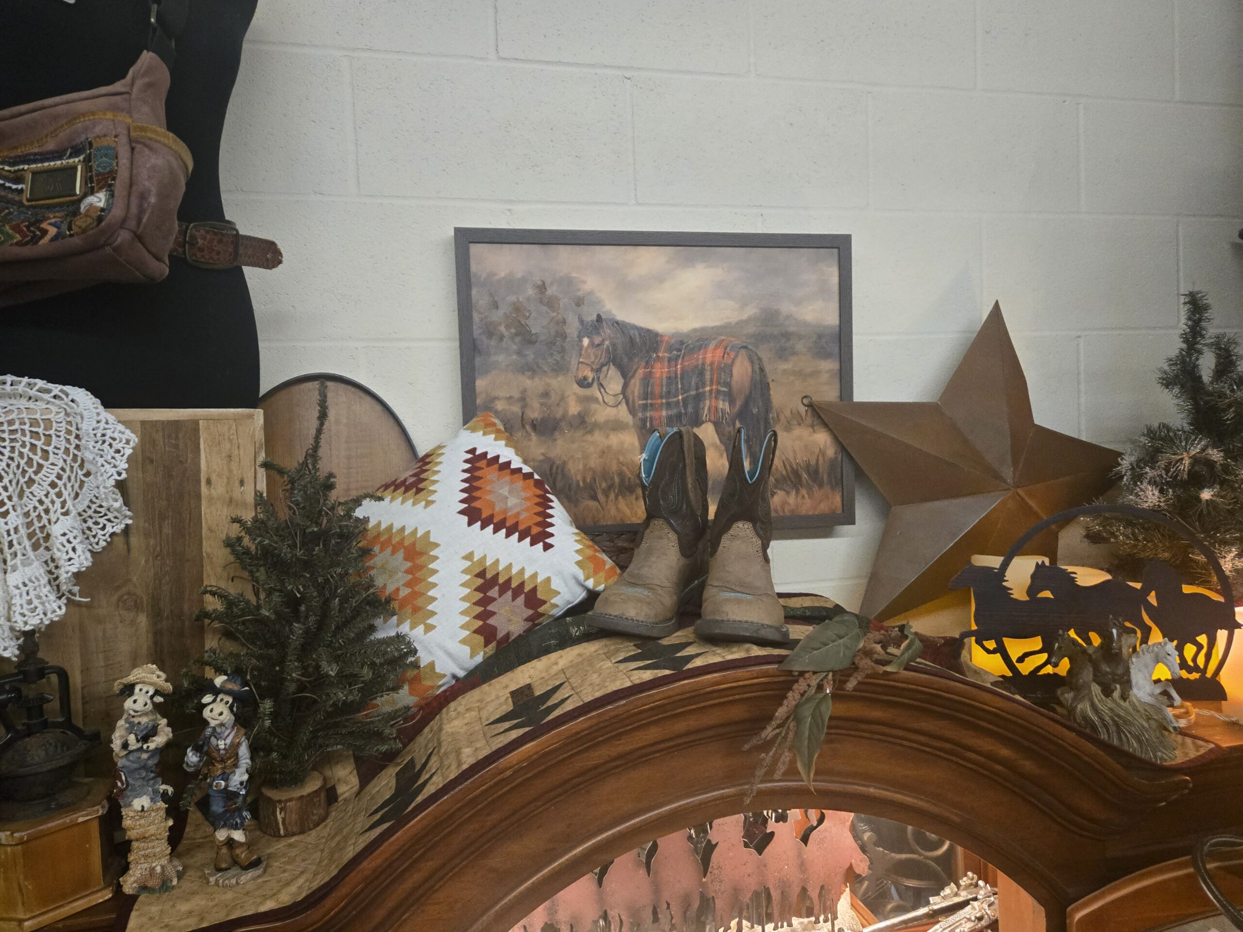 Framed painting of a bay horse wearing a plaid blanket on open grassland, a rust-brown metal barn star, children’s brown cowboy boots with turquoise lining, a Southwestern geometric throw pillow, a Southwestern-pattern quilt, a running-horses metal silhouette candle holder, resin horse figures, resin cow figurines in overalls and a Western vest, a cast-iron coffee grinder, a crocheted doily, a small evergreen tree on a log base, and a suede belt bag with a woven panel on a dress form at Shop 217, Pacific Antiques Mall in Tacoma, WA.