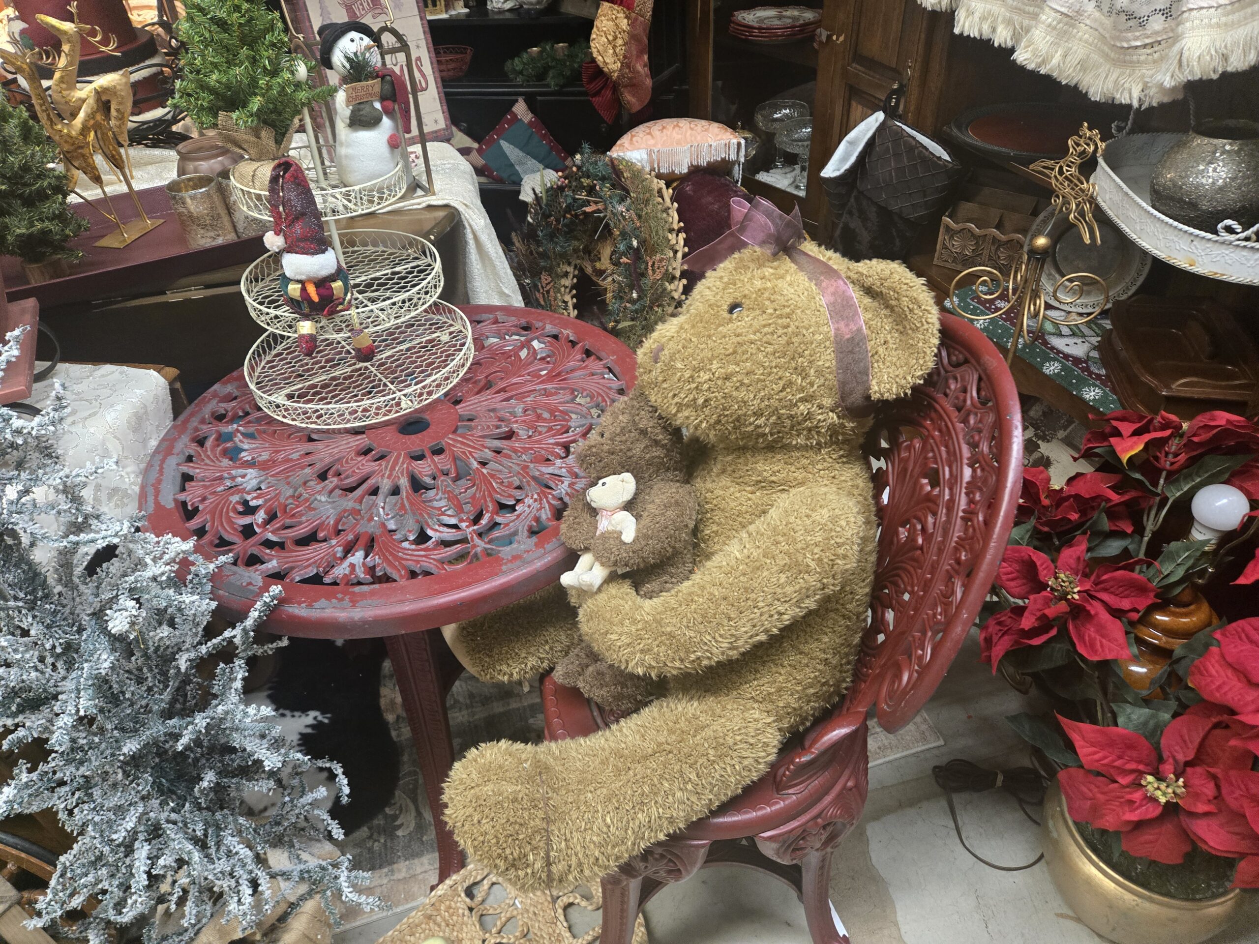 Oversized plush teddy bear with a pink ribbon holding a smaller brown bear and a cream bear, seated on a red cast-metal bistro chair beside a matching table with leaf scroll openwork, a tiered wire stand with a plush snowman in a knit hat, a flocked artificial tree, red faux poinsettias in a brass pot, a gold metal reindeer, a snowman holding a Merry Christmas sign, a velvet patchwork stocking, and a gold wire reindeer at Shop 217, Pacific Antiques Mall in Tacoma, WA.
