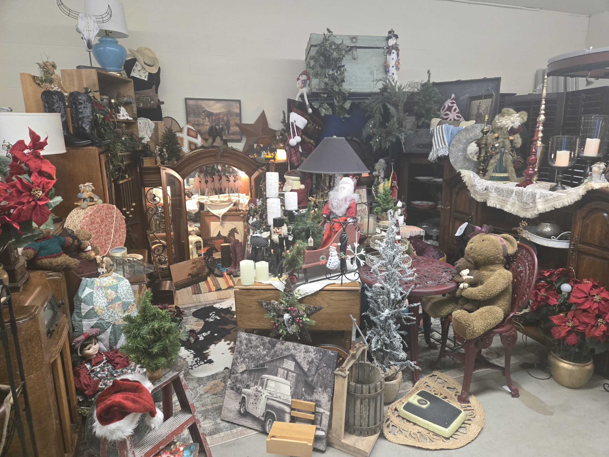 Western and Christmas shop with an arched wood china cabinet, a whitewashed steer skull sculpture, a blue ceramic table lamp, a straw cowboy hat on a dress form, tall black cowboy boots, a plaid-blanket horse painting, a metal barn star, a mint green metal trunk, plush snowmen, Santa figures, a red wooden sleigh with a plush Santa, a black nutcracker, an oak drop-leaf table, a red cast-metal bistro set with plush teddy bears, red faux poinsettias, a Silvertone console radio, a porcelain doll, a patchwork quilt, a floral fabric hatbox, a black-and-white pickup truck and barn canvas, a slatted wooden barrel press, a yellow bathroom scale, a cowhide rug, and a braided jute rug at Shop 217, Pacific Antiques Mall in Tacoma, WA.
