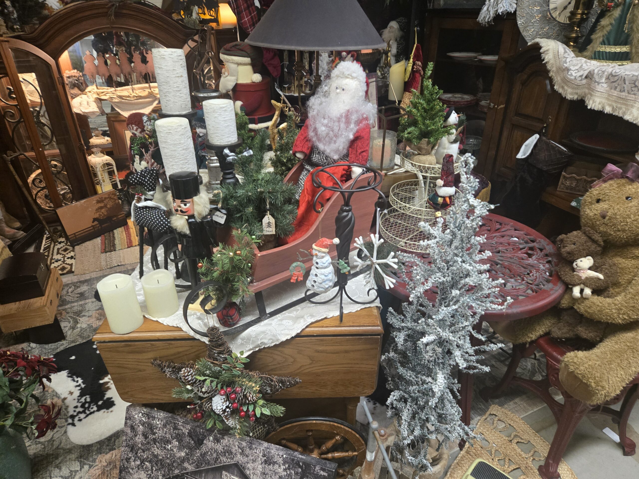 Red wooden sleigh with black metal runners holding a plush Santa with a curly white beard, a black wooden nutcracker with a fur beard, small artificial evergreen trees, white birch-texture LED pillar candles on black candlesticks, cream LED candles, a ceramic snowman ornament, a white snowflake, a wrought iron candle stand, an oak drop-leaf table, a twig star with pinecones and red berries, a flocked tree, a tiered wire stand, a red cast-metal bistro table, a spoked wooden wheel, and a cowhide rug at Shop 217, Pacific Antiques Mall in Tacoma, WA.