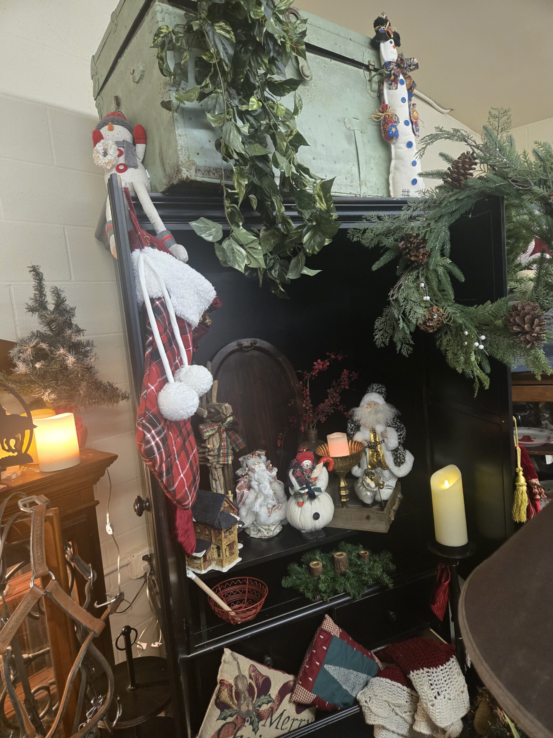 Black wood cabinet with drawers holding a white-robed Santa with a pink cape, a white and gold Santa with leopard-print trim, a snowman with a plaid scarf, a ceramic Christmas village house, a twig reindeer with a plaid bow, a brass pedestal candle bowl, LED pillar candles, a red wire basket, and an evergreen candle ring with log candle cups, with a red plaid stocking with sherpa cuff and pompoms, plush snowmen, a pinecone evergreen wreath, faux pothos vines, a mint green metal trunk, and needlepoint and patchwork Christmas pillows at Shop 217, Pacific Antiques Mall in Tacoma, WA.