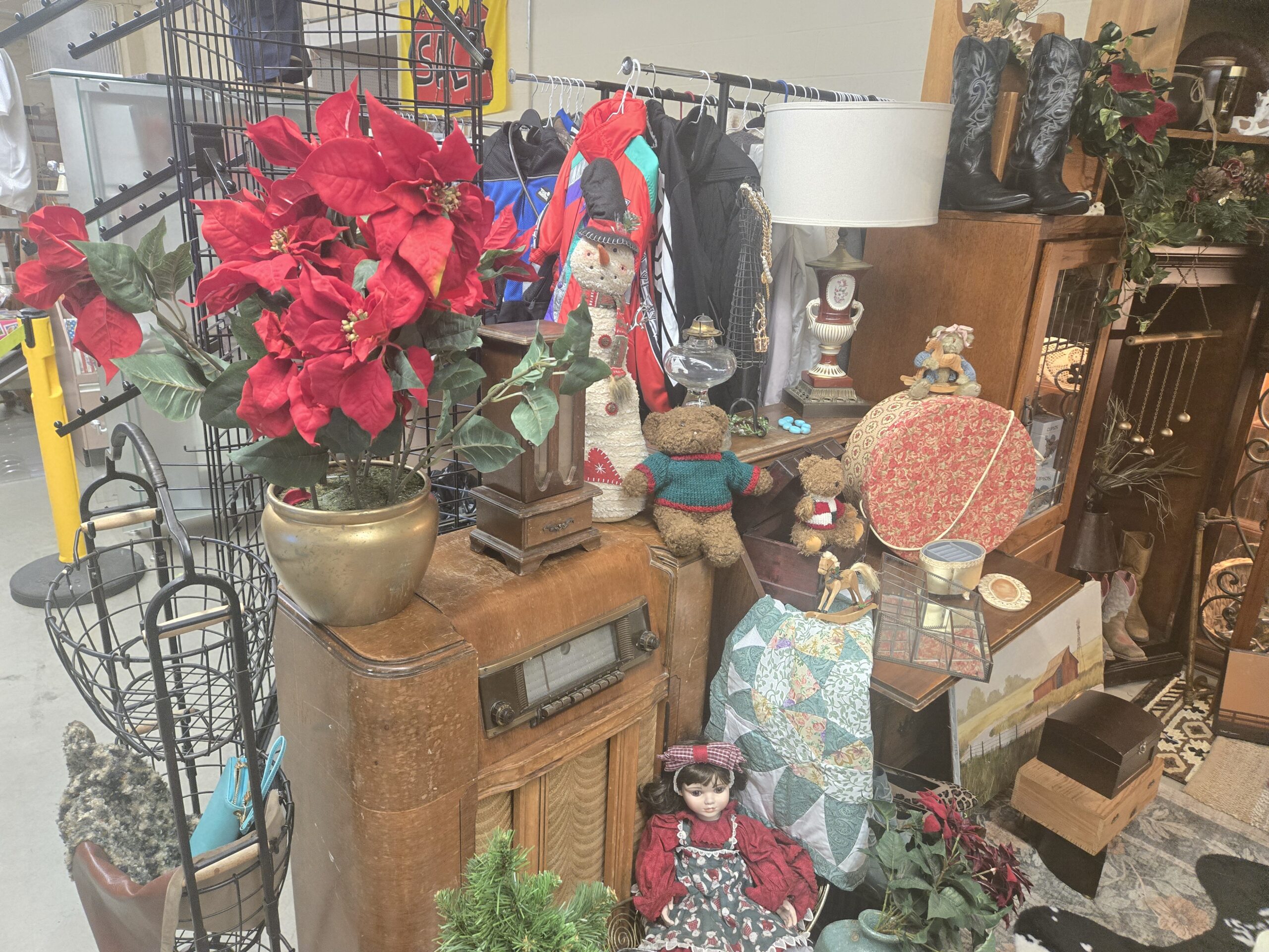 Silvertone console radio cabinet with a push-button dial, red faux poinsettias in a brass planter, a wooden tower box with a drawer, a fabric snowman, teddy bears in knit sweaters, a clear glass oil lamp, a porcelain urn lamp with a floral medallion, a wooden rocking horse ornament, a floral fabric hatbox, a beveled glass and brass box, a patchwork quilt, a porcelain doll in a red dress and print pinafore, a wire dress form necklace display, wire baskets, a red barn painting, and tall black cowboy boots at Shop 217, Pacific Antiques Mall in Tacoma, WA.