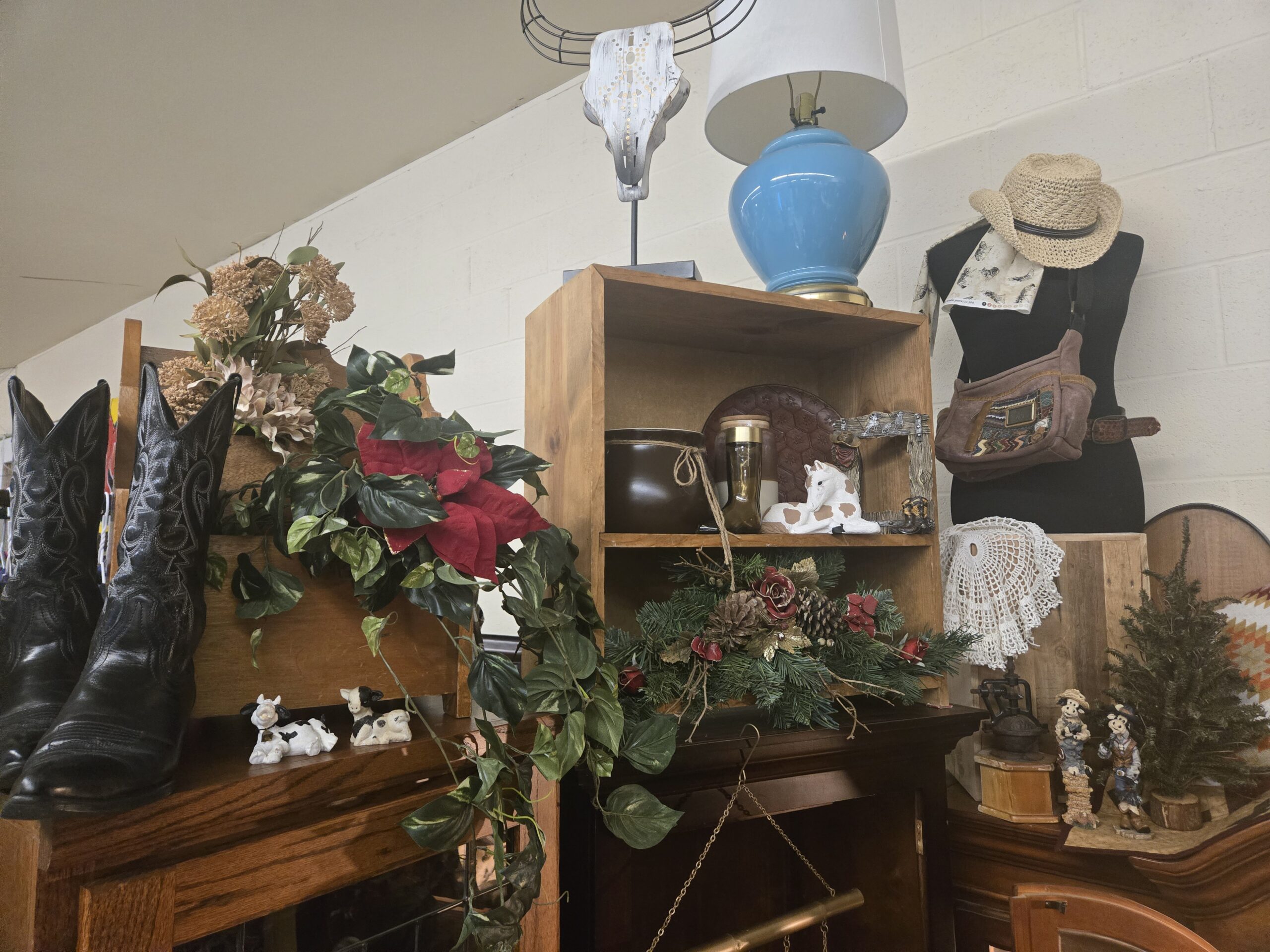 Tall black cowboy boots with white decorative stitching, a whitewashed steer skull sculpture with wire horns on a metal stand, a blue ceramic table lamp with a brass base, a wooden crate shelf, a brown glazed crock with twine, a brown glass boot-shaped jar, a resting pinto foal figurine, a rustic picture frame with cowboy boot and rope accents, black and white calf figurines, a wooden wall rack with dried florals and faux pothos, an evergreen centerpiece with red roses and pinecones, a straw cowboy hat and horse-print scarf on a dress form, a suede belt bag with a woven panel, a crocheted doily, a cast-iron coffee grinder, and resin cow figurines at Shop 217, Pacific Antiques Mall in Tacoma, WA.