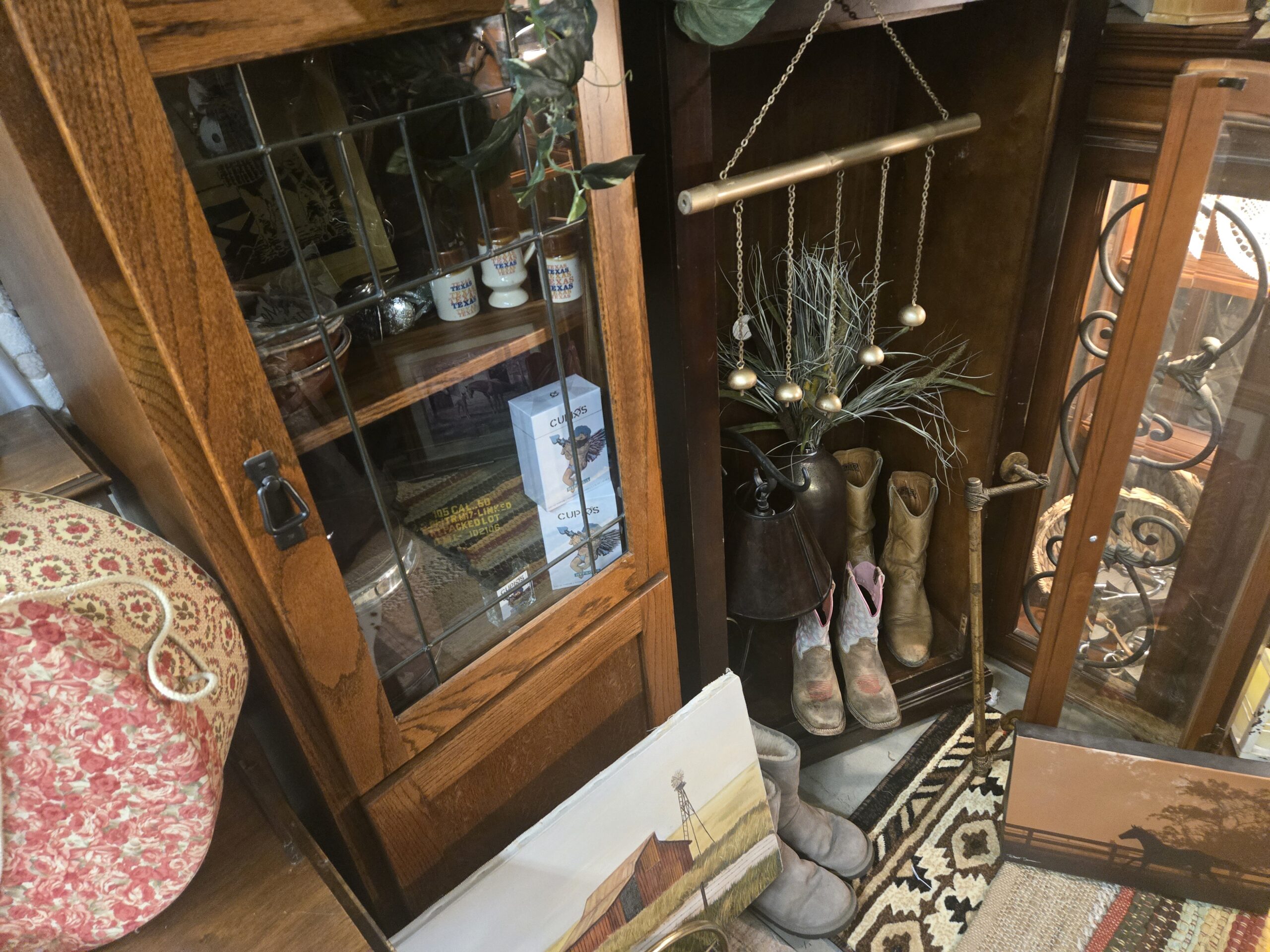 Oak cabinet with a leaded glass door holding three Texas souvenir mugs, boxed Cupid’s fragrance, a framed horse print, stacked brown bowls, and a silver ornament, beside tan leather cowboy boots, pink and tan children’s cowboy boots, a brass bar wind chime with gold bells and a grass spray, a leather lamp shade, gray suede boots, a barn and windmill painting on canvas, a horse silhouette fence-line print, a Southwestern rug, and a floral fabric hatbox at Shop 217, Pacific Antiques Mall in Tacoma, WA.