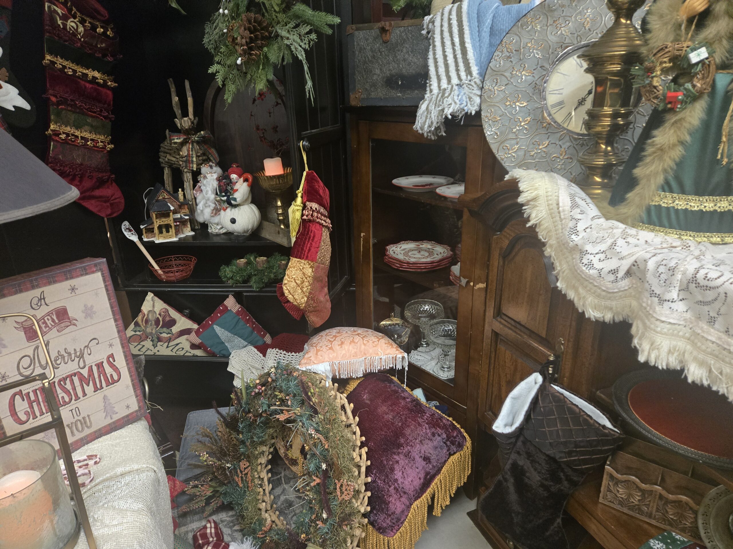 Velvet patchwork Christmas stockings with gold trim, an A Very Merry Christmas to You sign with a plaid border, a ceramic Christmas village house, a twig reindeer with a plaid bow, Santa and snowman figures, a brass pedestal candle bowl, red and cream scalloped plates, white plates with red bird and star motifs, diamond-pattern pressed glass compotes, a twig wreath with dried florals, a purple velvet pillow with gold fringe, a peach fringed pillow, a quilted brown stocking, needlepoint and patchwork Christmas pillows, and an embossed fleur-de-lis wall clock at Shop 217, Pacific Antiques Mall in Tacoma, WA.