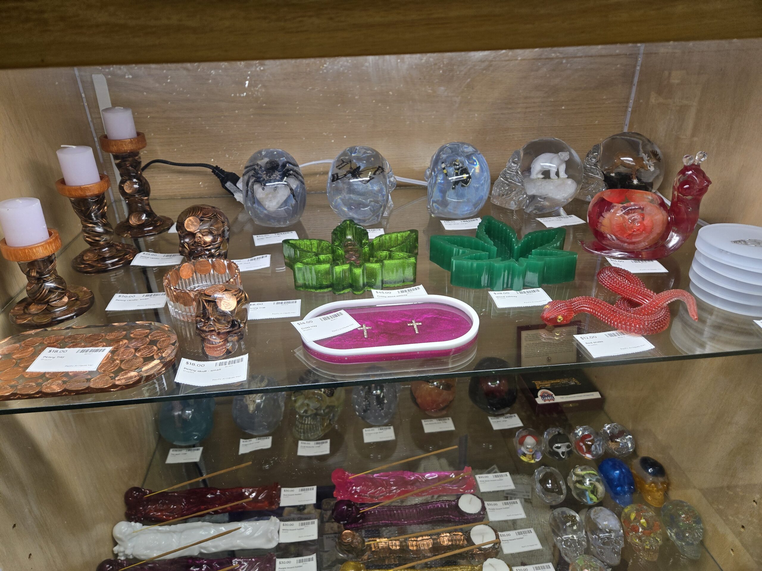Handmade clear resin skulls with a black spider on white stone, a winged insect, a black-and-yellow figure, a polar bear and a moose cast inside, copper penny resin candle holders with white pillar candles, small and large penny skulls, a penny cup, an oval penny tray, green glitter and green cannabis-leaf ashtrays, a pink glitter tray with two gold crosses, a red resin snail, a red resin snake, and skull-end resin incense holders on the shelf below at Shop 333, Pacific Antiques Mall in Tacoma, WA.