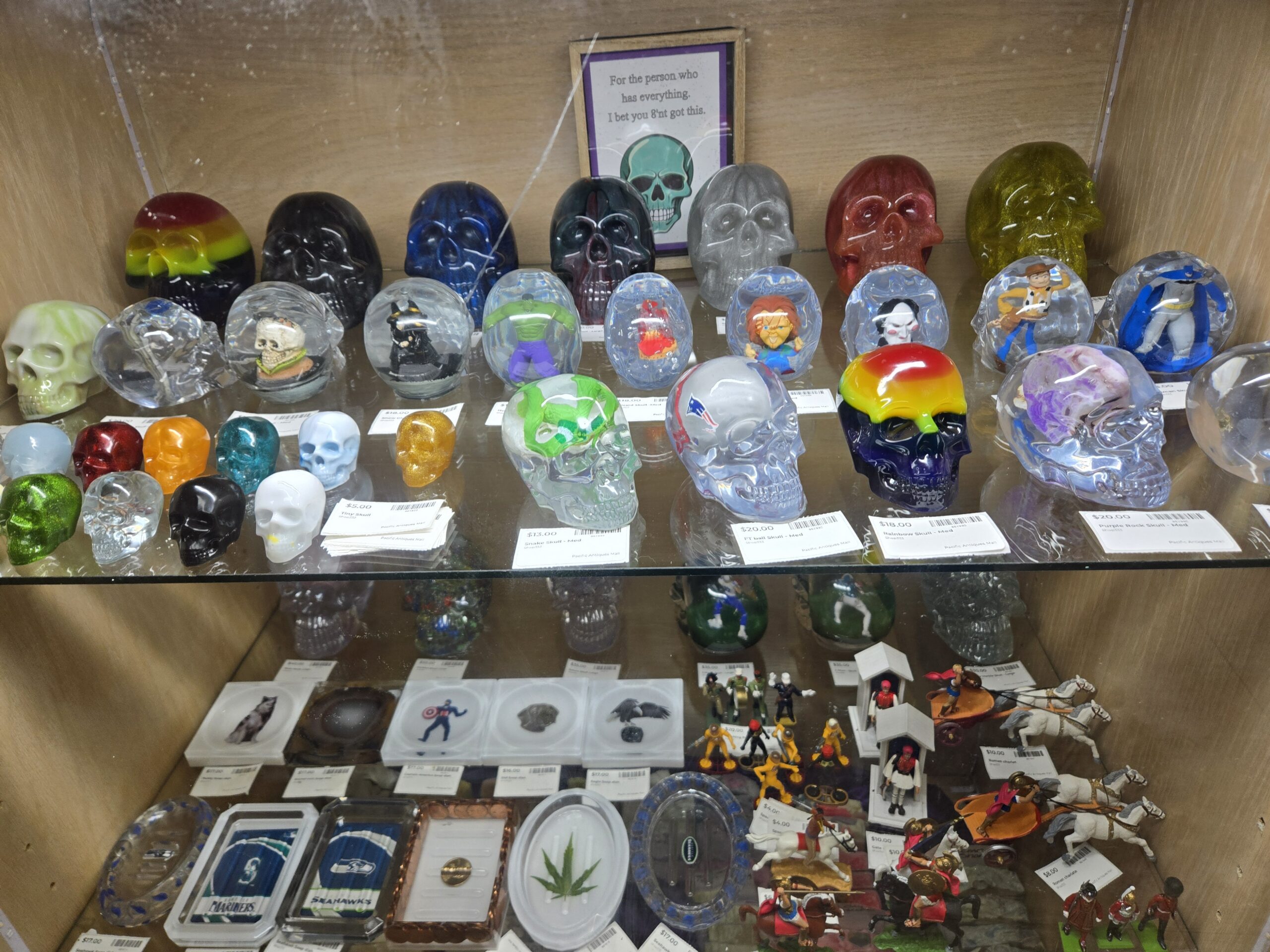 Solid resin skulls in rainbow stripes, black, cobalt blue, gray, red glitter and gold glitter, clear resin skulls enclosing Chucky, the Saw puppet, the Hulk, Woody from Toy Story and Batman, a green snake skull, a New England Patriots logo skull, a rainbow drip skull, a purple rock skull, tiny skulls in red, orange, teal, gold glitter, green glitter, black and white, a framed teal skull sign, and resin soap dishes with Seattle Mariners and Seahawks logos beside hand-painted Roman chariot and guard figures on the shelf below at Shop 333, Pacific Antiques Mall in Tacoma, WA.