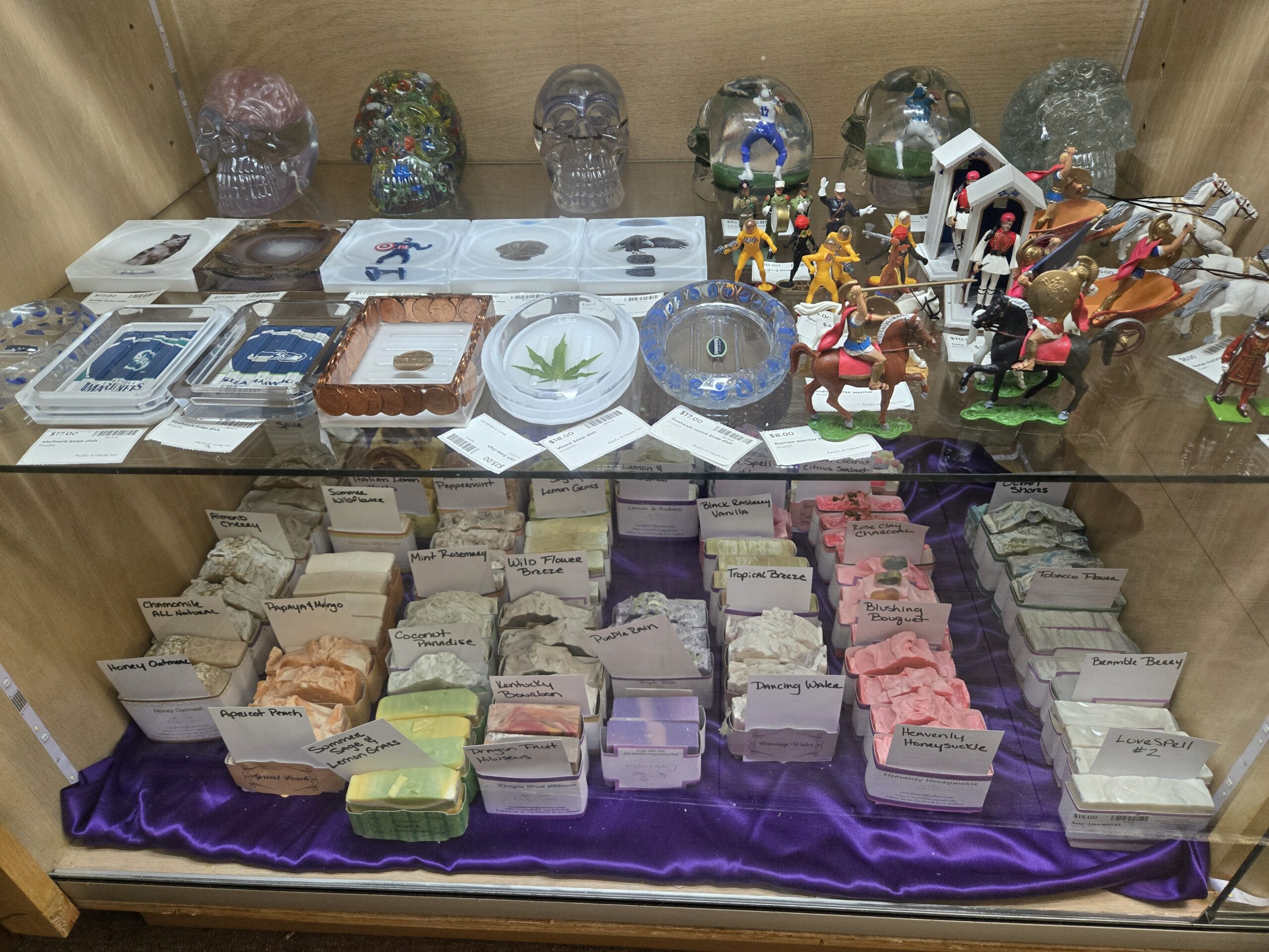 Resin soap dishes with a husky, Captain America, a bald eagle, a polished agate slice, a cannabis leaf, a copper penny border, Seattle Mariners and Seattle Seahawks logos and a round Seahawks dish, hand-painted plastic Roman chariots with white horses, mounted Roman warriors, Greek Evzone guards in white sentry boxes, yellow spacesuit figures, clear and confetti-color resin skulls, football player figures in resin domes, and Crafty Crafters bar soaps including Honey Oatmeal, Chamomile All Natural, Papaya & Mango, Kentucky Bourbon, Purple Rain, Dancing Water, Heavenly Honeysuckle, Bramble Berry and Love Spell #2 on purple satin at Shop 333, Pacific Antiques Mall in Tacoma, WA.