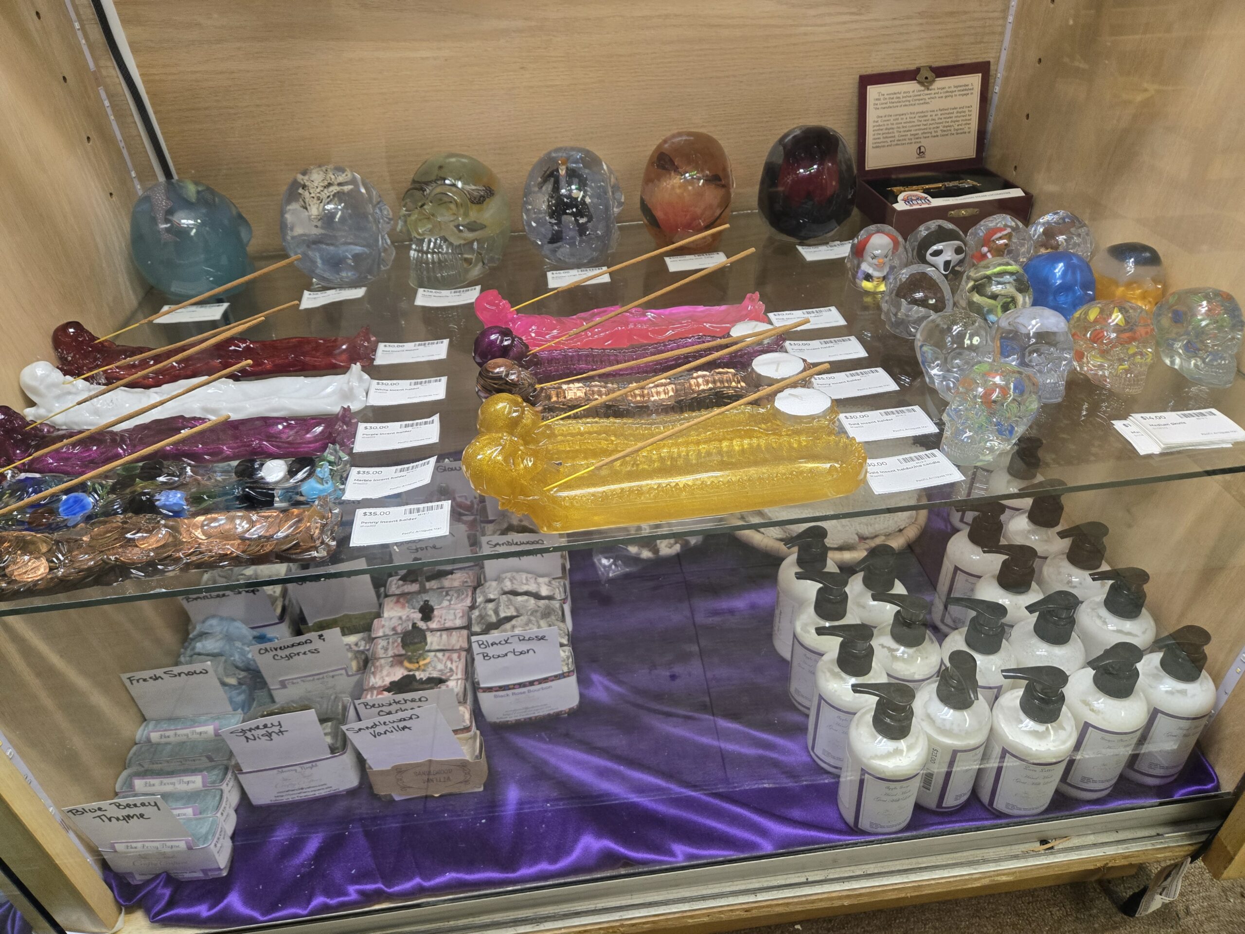 Resin skulls enclosing a dragon skull, butterflies and a figure in a black suit, skull-end incense holders in red, white, purple, pink, gold glitter, copper penny and marble-filled clear resin with tea light cups, small resin skulls with Pennywise and Ghostface figures, clear skulls with confetti-color inclusions, a Lionel Hudson steam locomotive miniature in a burgundy presentation box, Crafty Crafters bar soaps labeled Fresh Snow, Olive Wood and Cypress, Black Rose Bourbon and Starry Night, and handmade goat milk lotion in black pump bottles on purple satin at Shop 333, Pacific Antiques Mall in Tacoma, WA.