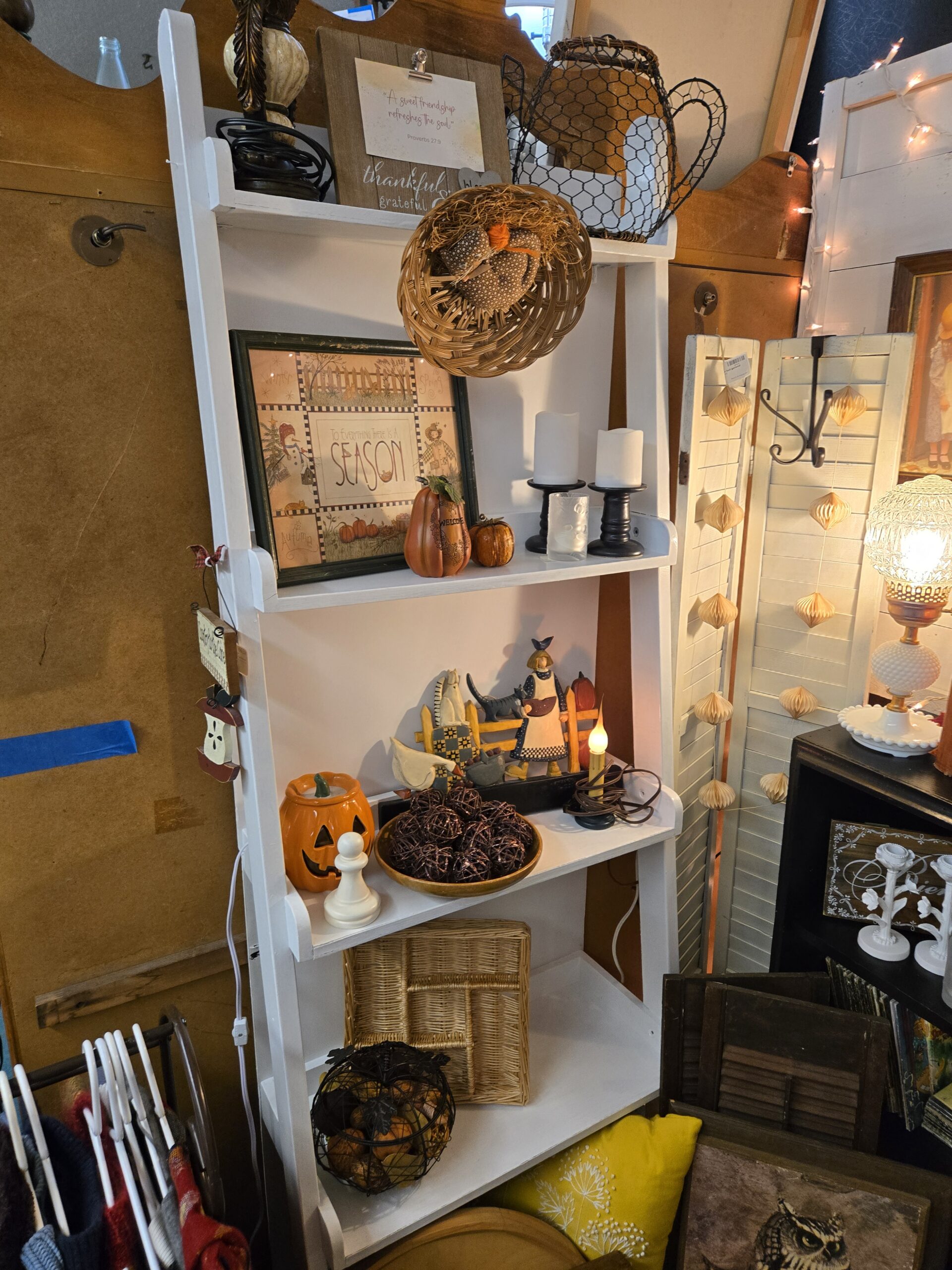 White ladder shelf with a framed To Everything There Is a Season folk-art print showing snowman, scarecrow and pumpkin patch panels, a chicken-wire teapot basket, a thankful grateful wood clipboard sign holding a Proverbs 27:9 card, a wicker basket with a fabric pumpkin, a ceramic Welcome pumpkin, flameless pillar candles on black turned-wood holders, a folk-art farm woman shelf sitter with a crow, cat and goose, a ceramic jack-o'-lantern, a wooden bowl of twig balls, an electric candle lamp, a wire pumpkin basket of faux gourds, and a Country to the Core hanging sign, beside white louvered shutters with a paper lantern garland and a hobnail milk glass lamp at Shop 53, Pacific Antiques Mall in Tacoma, WA.