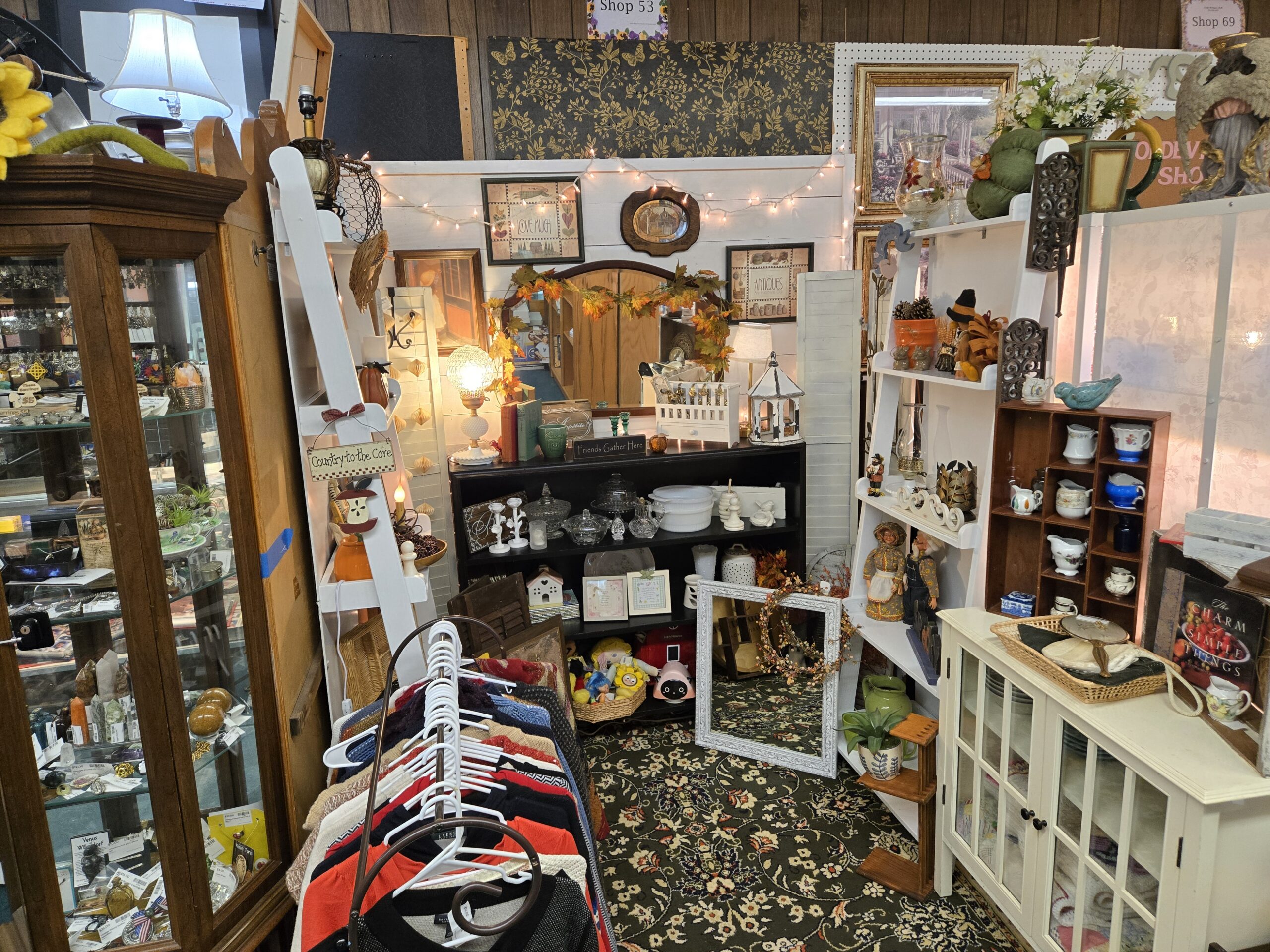 Two white ladder shelves of fall and Thanksgiving decor, a hobnail milk glass lamp with a pressed glass globe shade, a black bookcase of clear pressed glass and milk glass, framed folk-art prints, an oval framed print, an arched mirror draped in autumn leaf garland, a white wooden lantern, white louvered shutters with string lights, a wooden cubby shelf of porcelain creamers, a cream glass-door cabinet, an ornate white frame with a berry wreath, and a rack of knit sweaters on a floral rug at Shop 53, Pacific Antiques Mall in Tacoma, WA.