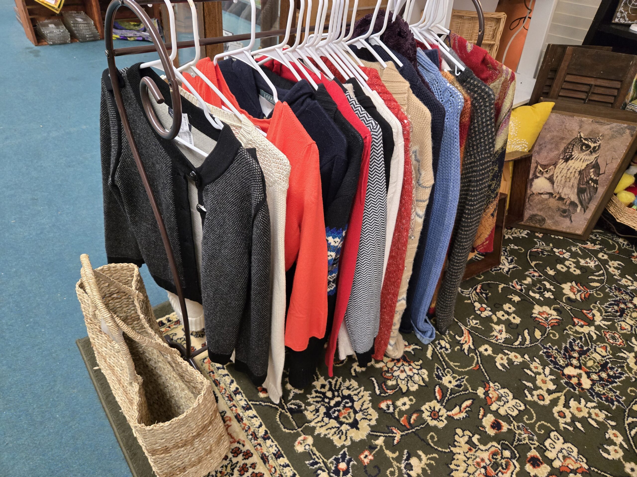 Rack of women's knit sweaters and cardigans including Lauren Ralph Lauren and Banana Republic labels, in charcoal bird's-eye knit, chunky cream knit, coral, navy, red, black-and-white chevron, oatmeal, blue open-knit, gray textured knit and a red patterned knit, with a woven seagrass tote bag and an owl print on wood board at Shop 53, Pacific Antiques Mall in Tacoma, WA.