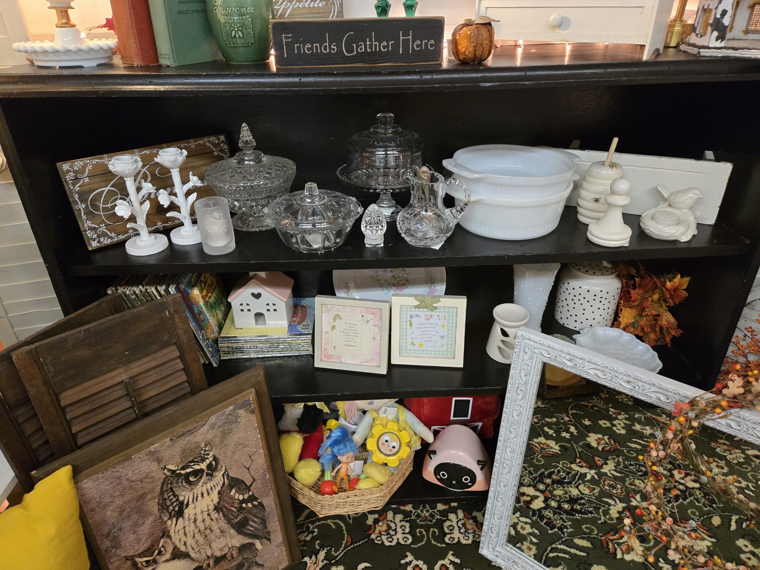 Black bookcase with a diamond-pattern pressed glass covered candy dish on a pedestal, a beaded-rim covered glass dish, a domed glass cake stand, a cut glass pitcher holding silver spoons, a cut crystal egg, white milk glass casseroles with tab handles, a ceramic beehive honey pot with a wooden dipper, white floral candle holders, a hobnail milk glass vase, a white ceramic tea light house with a pink roof, framed verse prints, children's storybooks with gold foil spines including a Donald Duck title, a basket of vintage toys with a blue-haired doll, a Grace Livingston Hill hardcover, a green Provence canister, a Friends Gather Here wood sign, and an owl print on wood board at Shop 53, Pacific Antiques Mall in Tacoma, WA.
