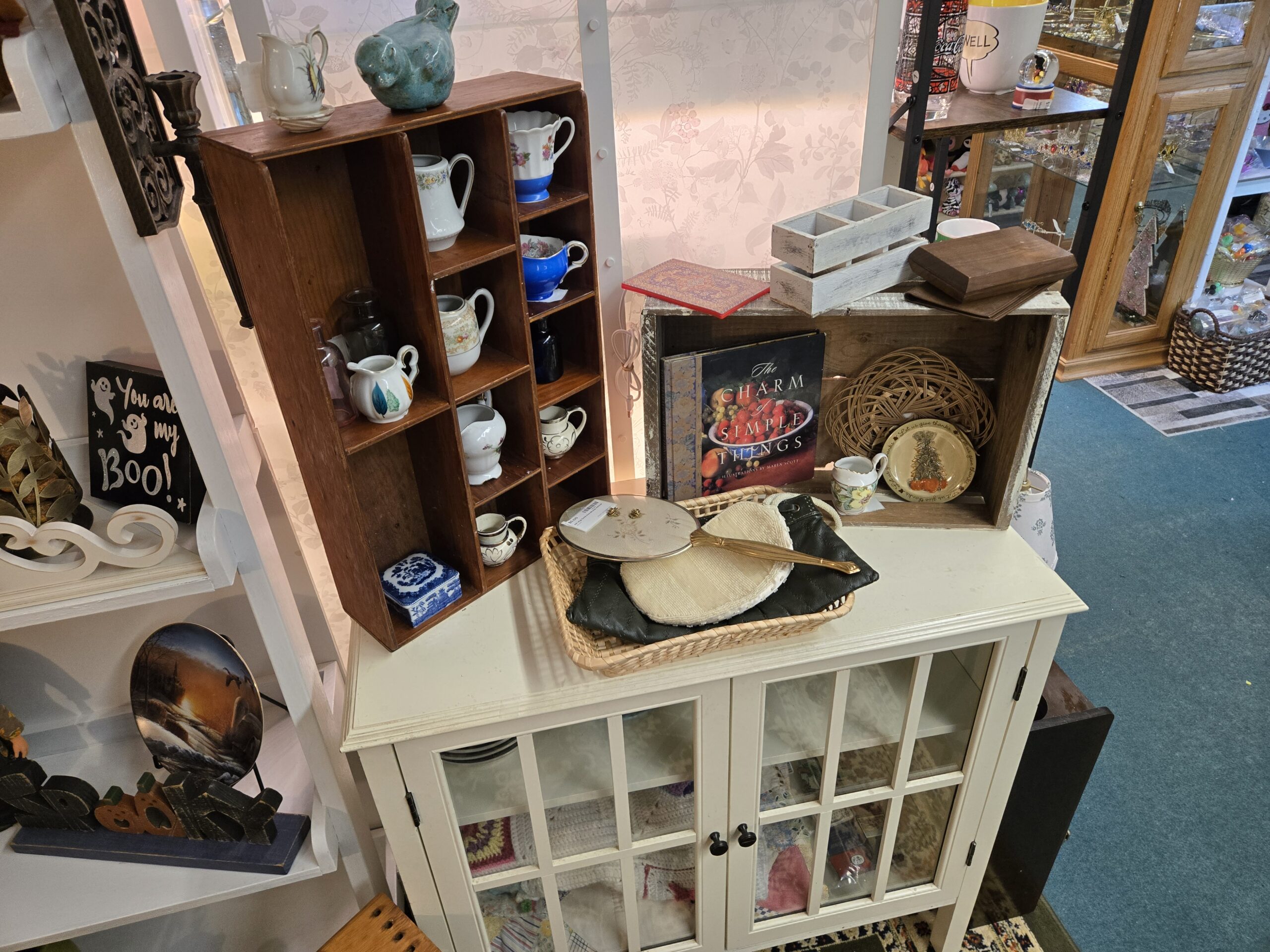Wooden cubby shelf of porcelain creamers including floral patterns, a cobalt blue creamer with a chintz interior, a cobalt-band creamer with a scalloped rim, a leaf-painted creamer and two silver lustre creamers, with a blue-and-white pagoda-scene trinket box and cobalt and pink glass bottles, a turquoise glazed ceramic bird, a wooden crate holding The Charm of Simple Things book illustrated by Maren Scott, a Let Us Give Thanks plate and a floral creamer, a wicker tray with a gold-tone vanity hand mirror with an embroidered floral back, a beaded cream clutch and a dark green quilted clutch, whitewashed wood caddies, and a cream glass-door cabinet holding crocheted afghans at Shop 53, Pacific Antiques Mall in Tacoma, WA.