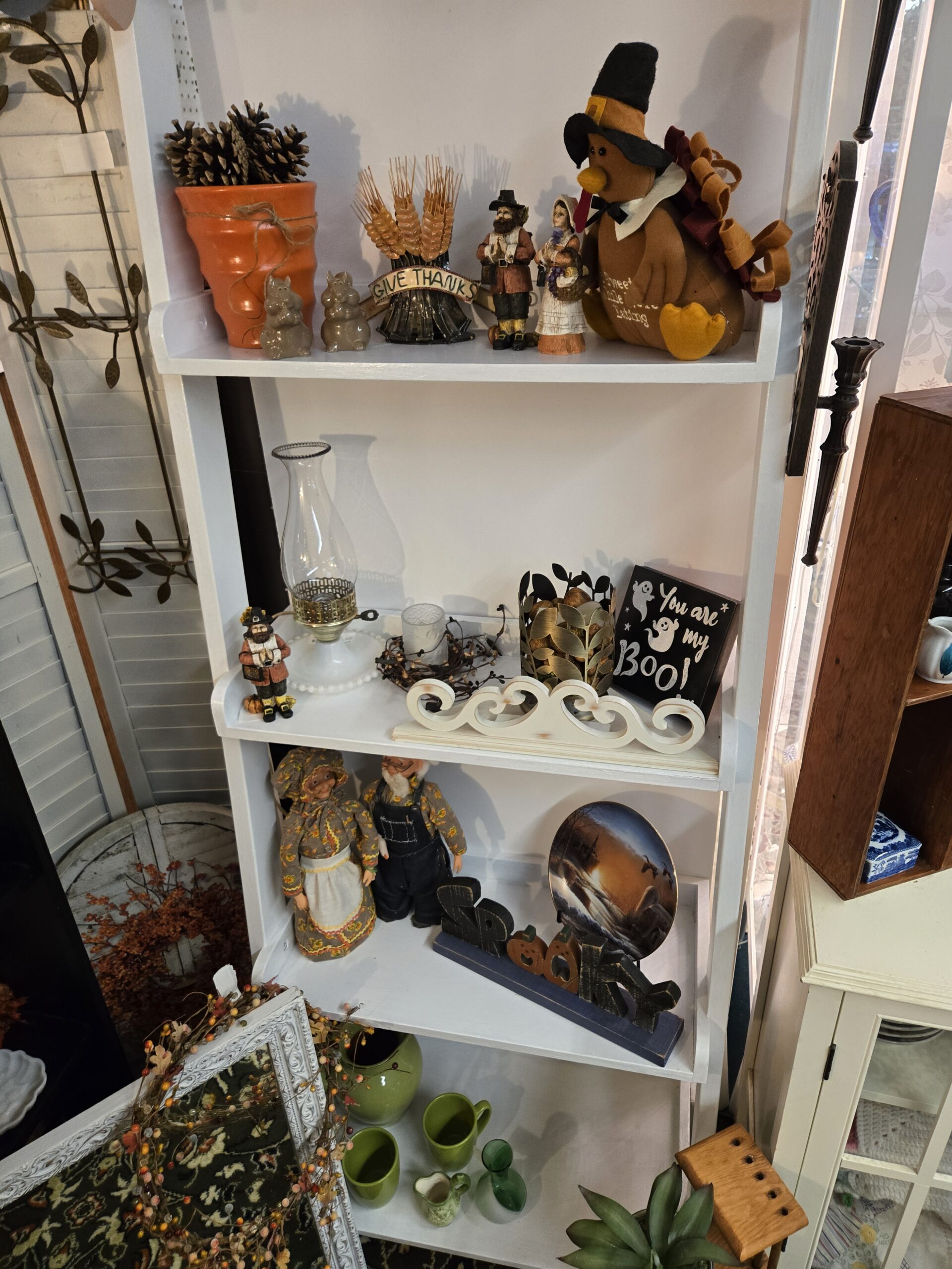 White ladder shelf with Pilgrim man and woman figurines, a plush turkey in a black buckled Pilgrim hat, a Give Thanks wheat-sheaf sign, a terracotta pot of pinecones, a pair of small glazed ceramic animal figurines, a hurricane oil lamp with a clear chimney and beaded milk glass base, a berry candle ring with a frosted votive, a gold-tone metal leaf candle holder, a You are my Boo! ghost sign, a white scrollwork shelf piece, calico-dressed grandma and grandpa dolls in an apron and denim overalls, a SPOOKY word sign with jack-o'-lantern letters, a collector plate of geese in flight over a sunset, and green ceramic mugs, a green pitcher and a green glass bottle at Shop 53, Pacific Antiques Mall in Tacoma, WA.