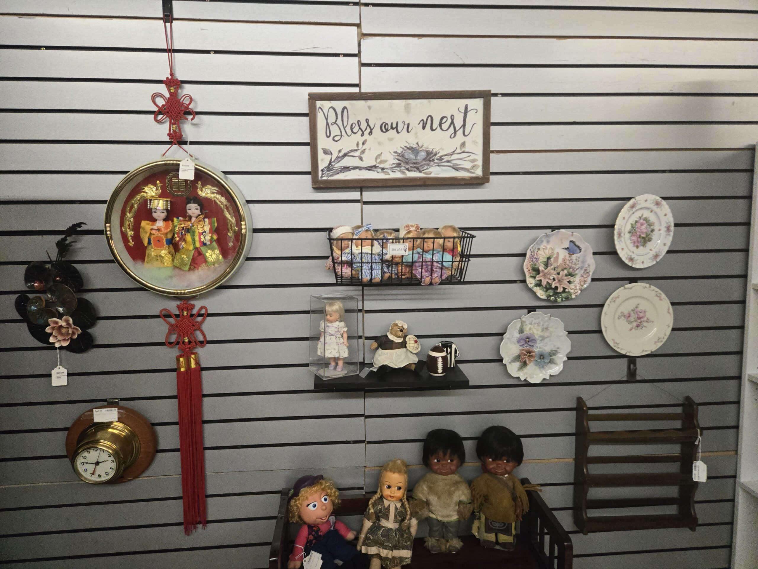 Round framed shadow box of bride and groom dolls in traditional Asian wedding dress with gilt phoenixes and red knotted tassels, a black metal lotus wall sculpture with a porcelain blossom, a brass ship’s clock on a round wood plaque, a “Bless our nest” sign, a wire basket holding a set of six baby dolls, a blonde doll in a lavender dress in a clear case, a teddy bear in a maid’s apron with a miniature football, four floral collector plates with roses, lilies, a butterfly and pansies, a wooden wall shelf, and a bench of dolls including a yarn-haired cloth doll, a braided-hair doll in a print dress, and two dolls in fur-trimmed outfits at Shop 54, Pacific Antiques Mall in Tacoma, WA.