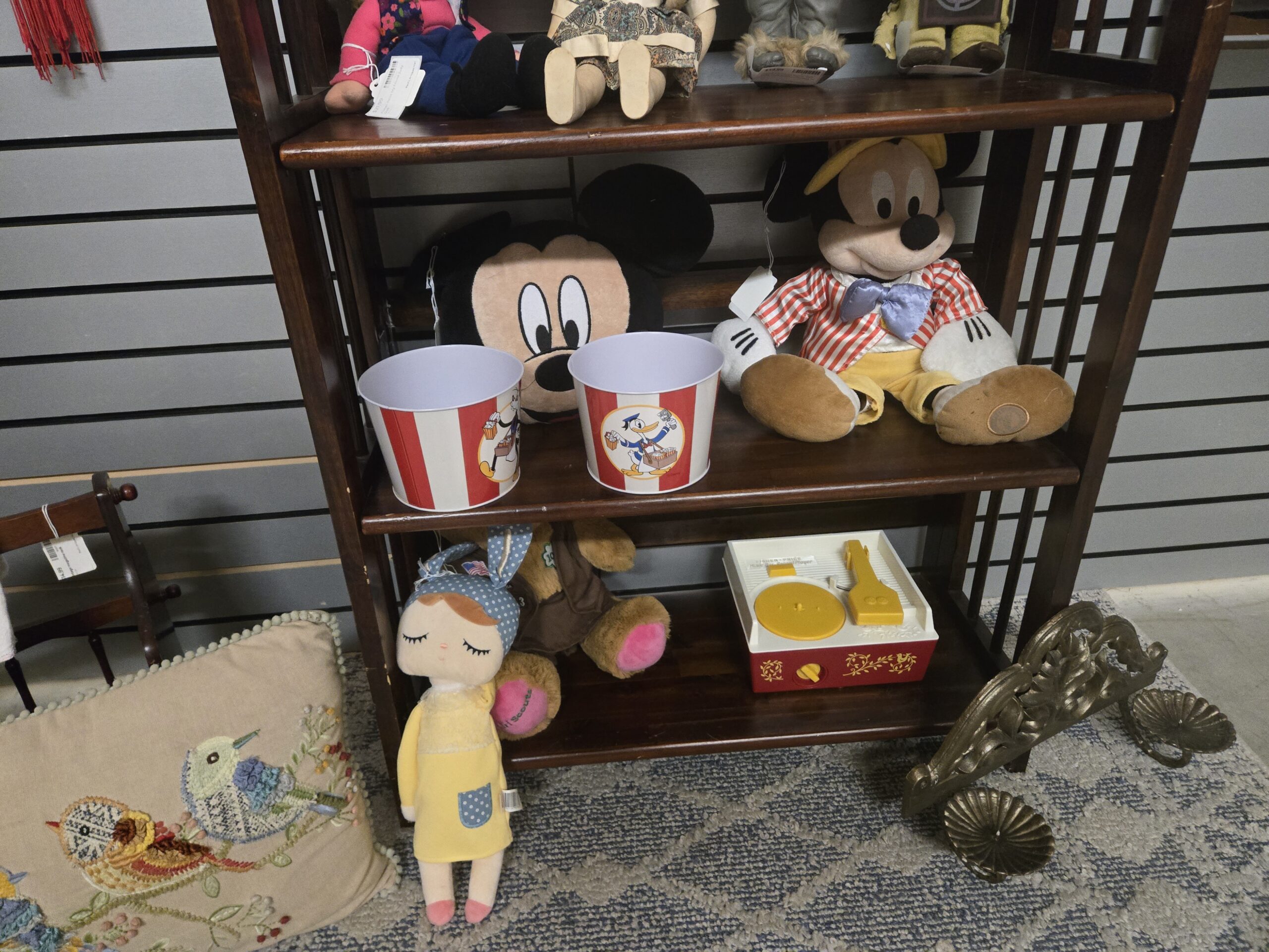 Two Mickey Mouse plush pieces, one an embroidered Mickey head pillow and one in a striped jacket and satin bow tie, Disney popcorn tins with Mickey Mouse and Donald Duck popcorn vendors, a Fisher-Price music box record player, a Girl Scouts teddy bear in a brown vest, a plush doll with a polka-dot bunny-ear hood, an embroidered bird pillow, a small wooden doll chair, and an ornate cast brass wall bracket with shell-shaped cups at Shop 54, Pacific Antiques Mall in Tacoma, WA.