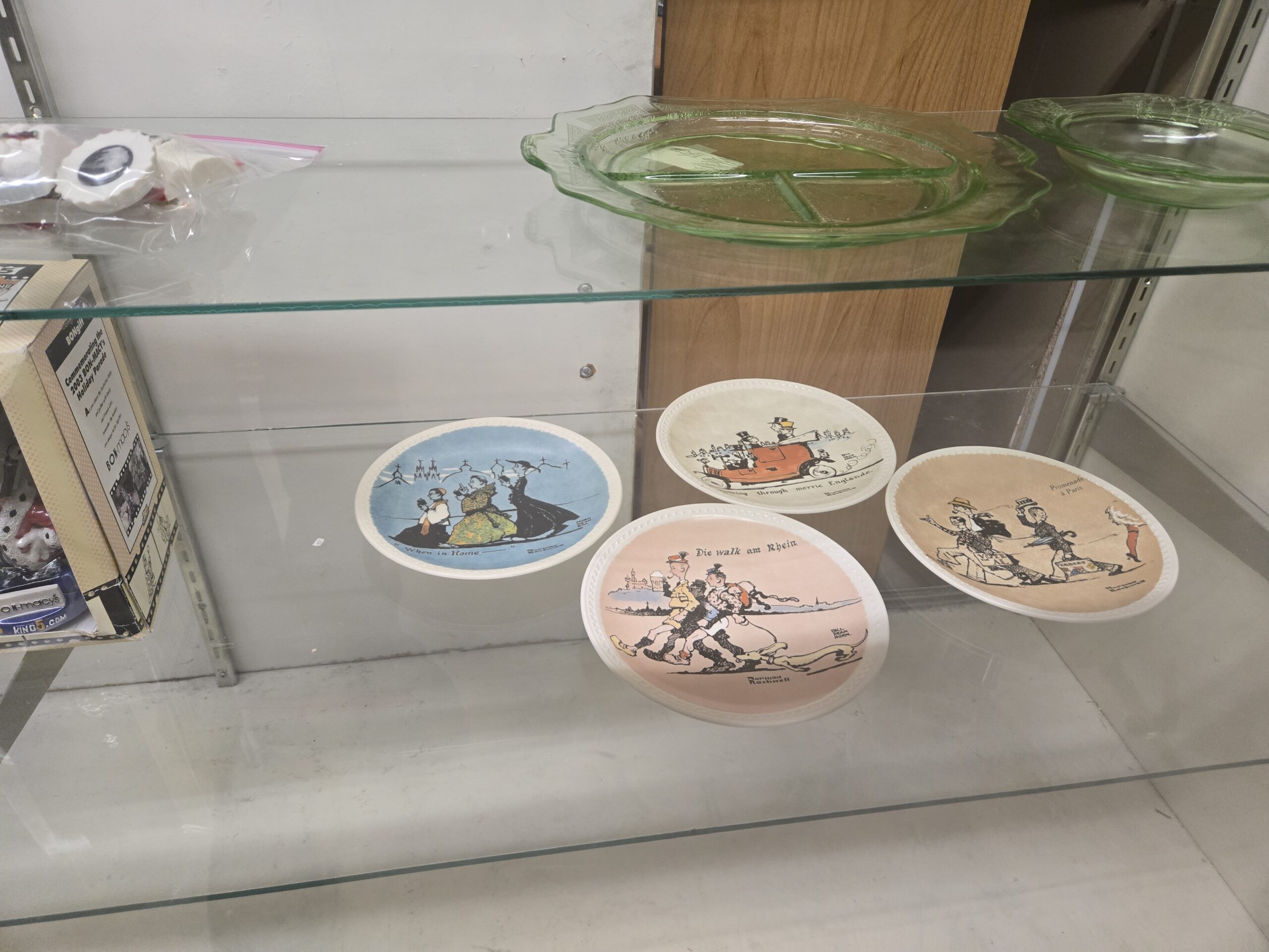 Four Norman Rockwell collector plates with European travel sketches titled “When in Rome,” “Die walk am Rhein,” “Promenade à Paris” and a Merrie Englande motoring scene, a green Depression glass grill plate, a second green Depression glass plate, and a boxed 2003 Bon-Macy’s Holiday Parade commemorative at Shop 54, Pacific Antiques Mall in Tacoma, WA.