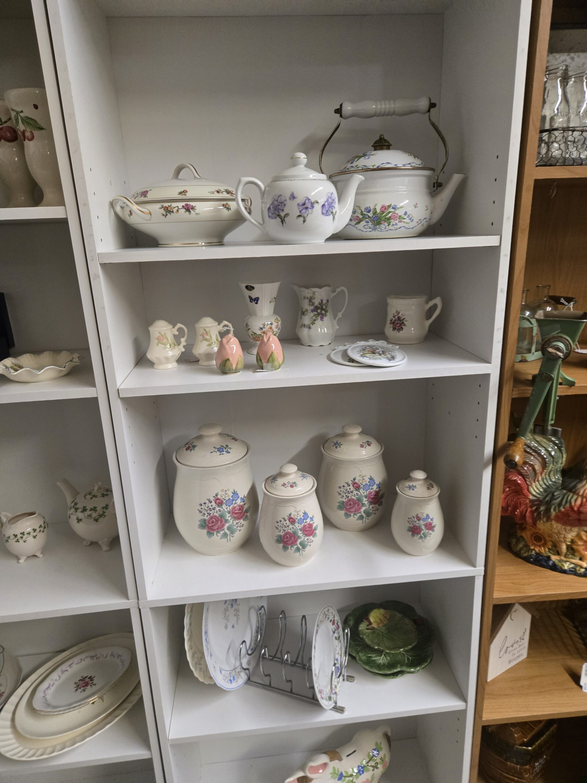 Gold-trimmed covered vegetable dish with scattered florals, a violet-painted teapot, a floral enamel tea kettle with porcelain handle and brass knob, tulip and rosebud salt and pepper shakers, a bone china vase with butterfly and floral decoration, a violet-painted pitcher, a rose-decorated mug, floral trivets, a four-piece canister set with pink roses and blue flowers, blue floral plates on a chrome plate rack, green cabbage-leaf plates, and a floral pig bank at Shop 54, Pacific Antiques Mall in Tacoma, WA.