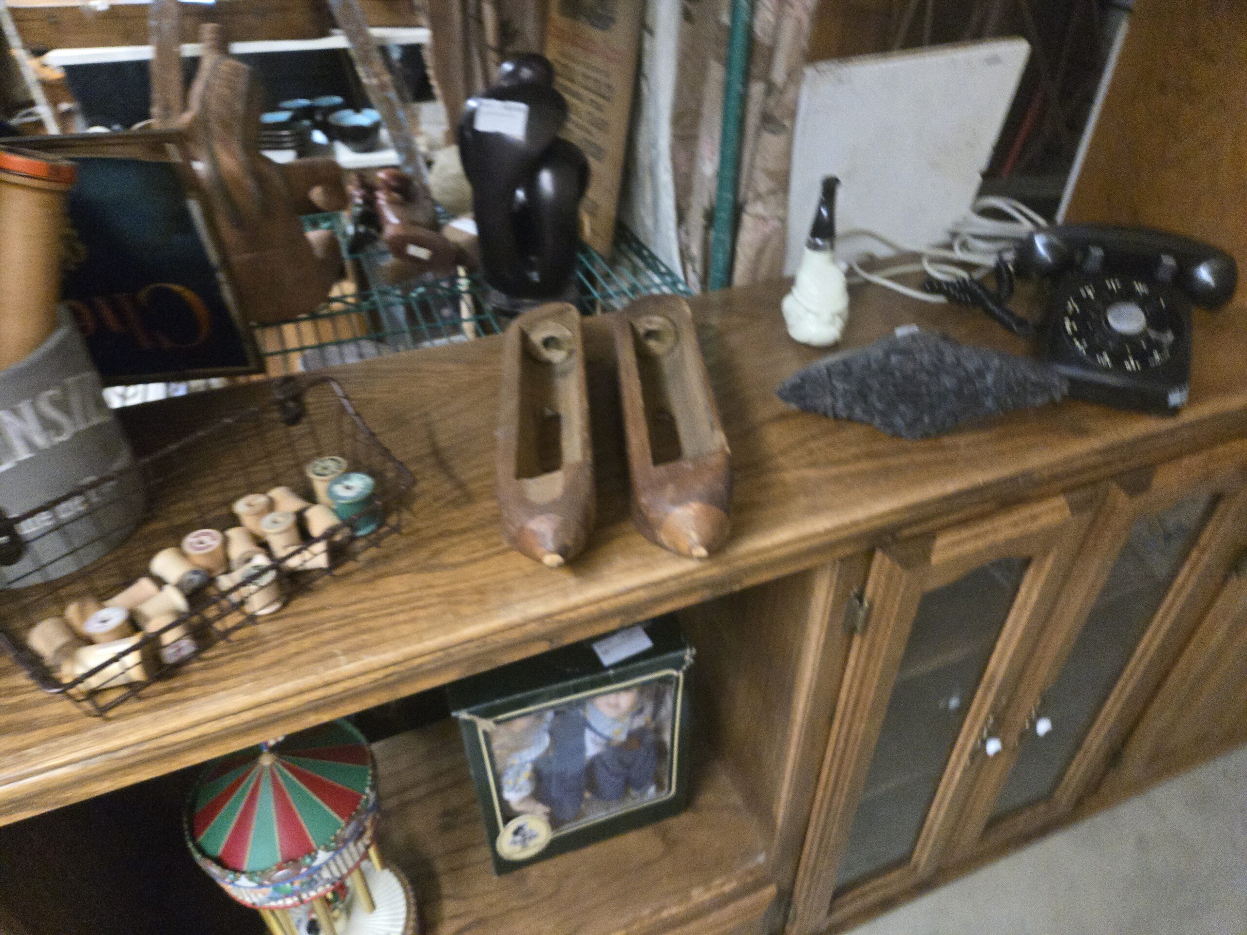 Wire basket of wooden thread spools, a pair of carved wooden shoe forms, a black carved figure, a white figural carving, a gray textured leaf-shaped piece, and a black rotary dial telephone, with a boxed Geppeddo doll pair and a carousel chime on the shelf below at Shop 54, Pacific Antiques Mall in Tacoma, WA.