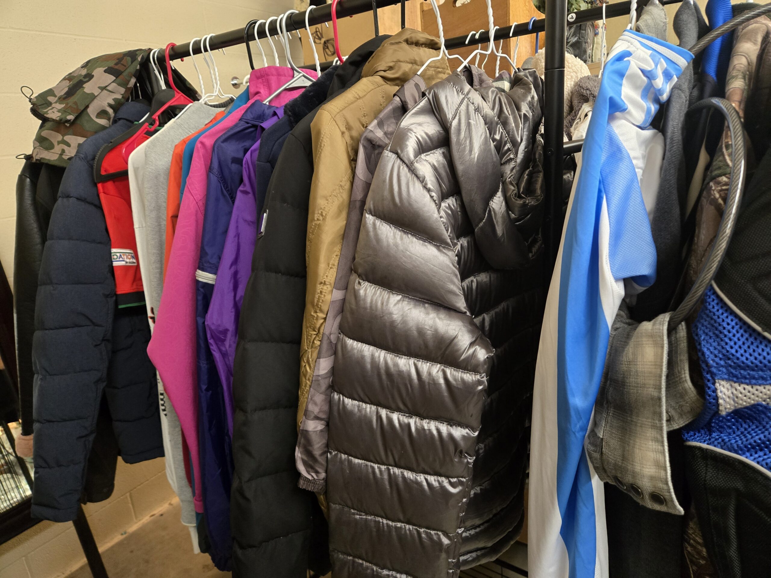 Woodland camo hooded jacket with drawcords, a navy quilted puffer, a red and black zip jacket with a sleeve patch, a gray sweatshirt, an orange jacket, a magenta soft-shell jacket with an embossed pattern, purple and violet nylon windbreakers, a long black puffer coat, an olive-gold nylon jacket, a gunmetal silver hooded puffer with gray camo-print sleeves, a blue and white athletic track jacket, a gray plaid shirt, and part of a blue Tour Master mesh motorcycle jacket at Shop 89, Pacific Antiques Mall in Tacoma, WA.