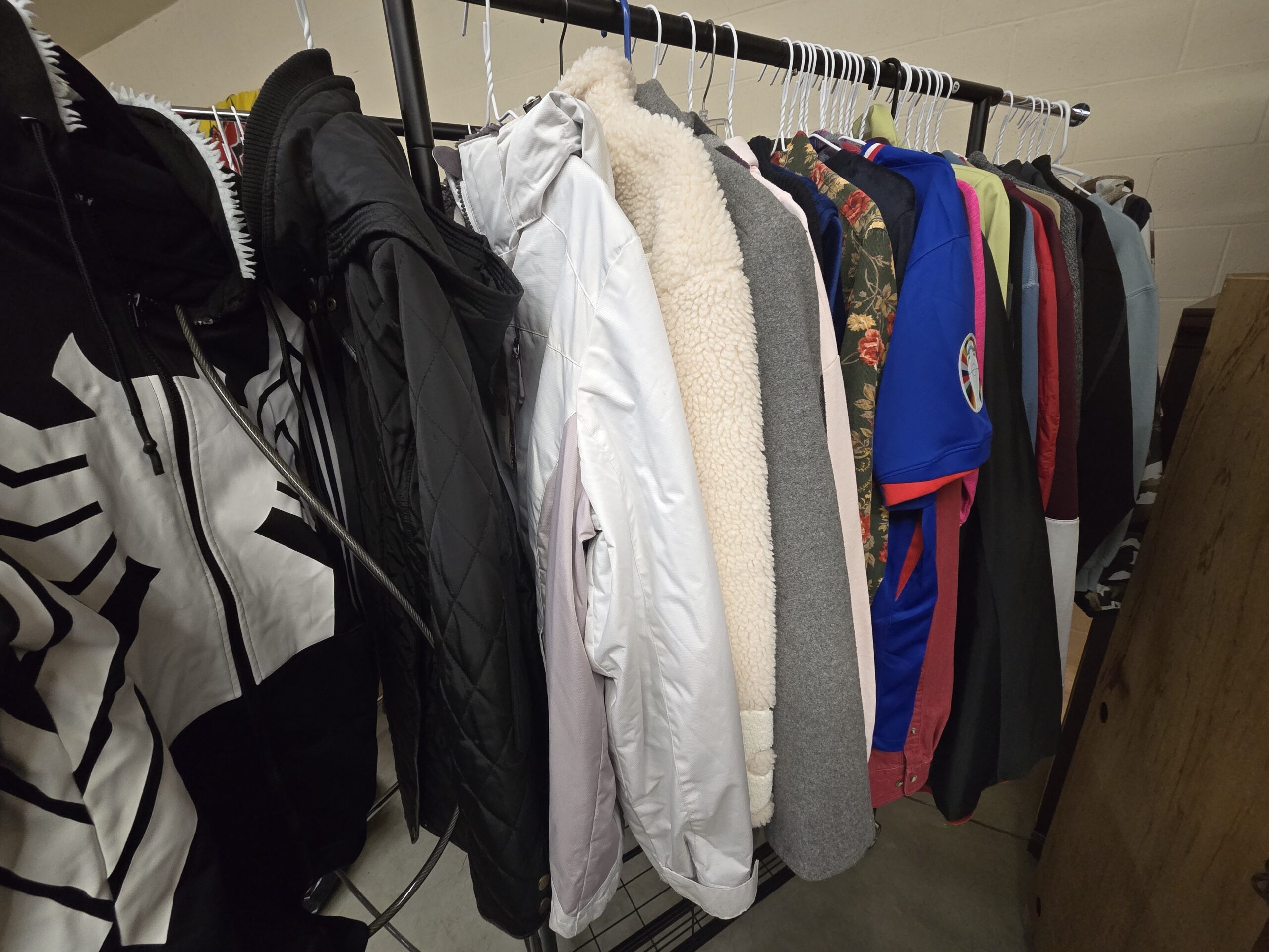 Black and white Venom-style zip hoodie with a fanged hood trim, a black diamond-quilted jacket with a knit collar and hood, a white and pale lilac insulated snow jacket, a cream sherpa fleece jacket, a gray long coat, a pale pink jacket, a green corduroy shirt with a red rose print, a blue soccer jersey with a UEFA Euro 2024 sleeve patch, and sweatshirts and shirts in red, maroon, lime, and light blue at Shop 89, Pacific Antiques Mall in Tacoma, WA.