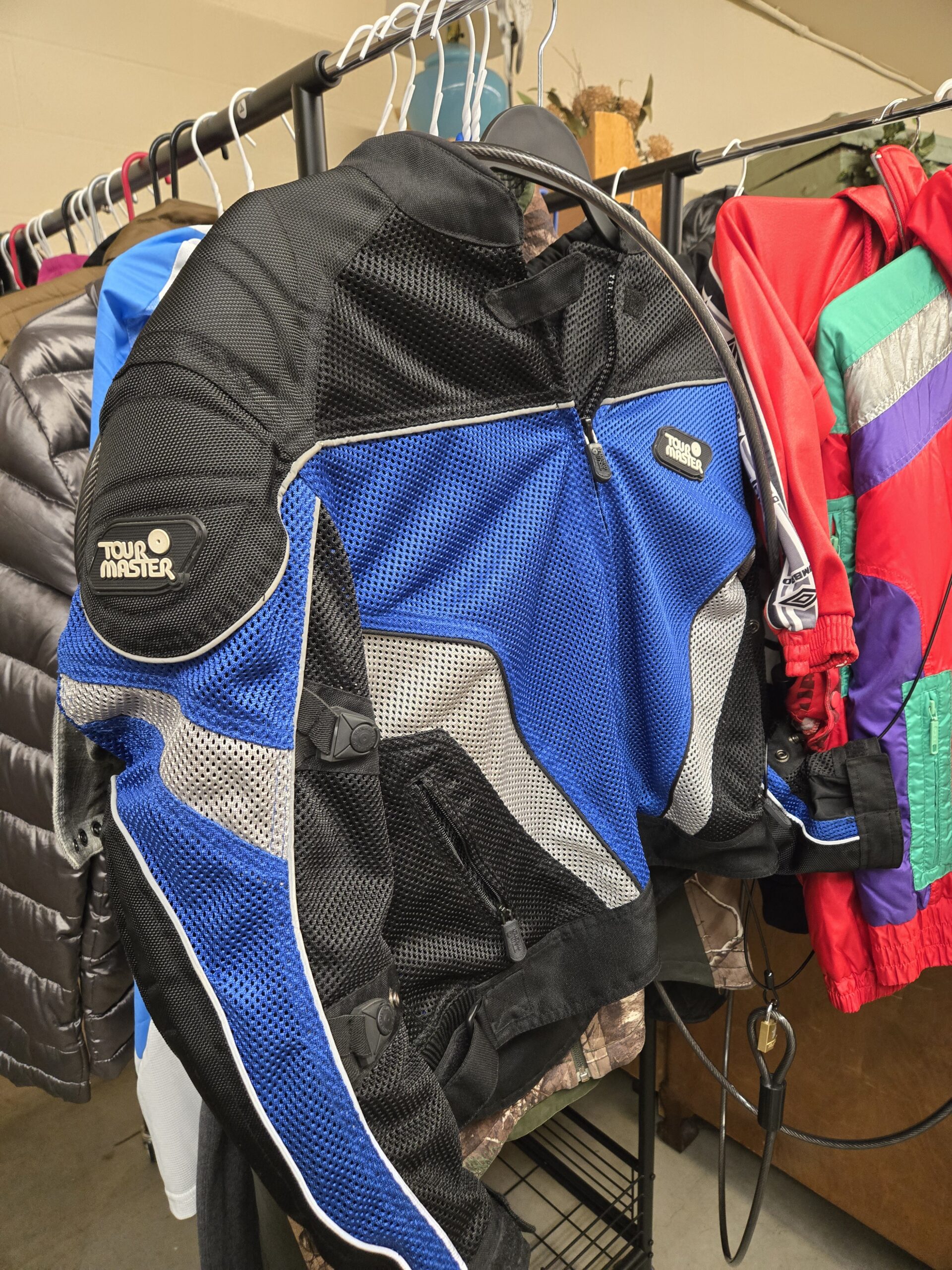 Tour Master mesh motorcycle jacket in blue, black, and silver with padded shoulder armor, Tour Master logo patches on the shoulder and chest, reflective piping, and adjustable side straps, hung beside a red Umbro track jacket, a vintage Spyder ski jacket, a gunmetal silver puffer, and a leaf-pattern camo jacket at Shop 89, Pacific Antiques Mall in Tacoma, WA.