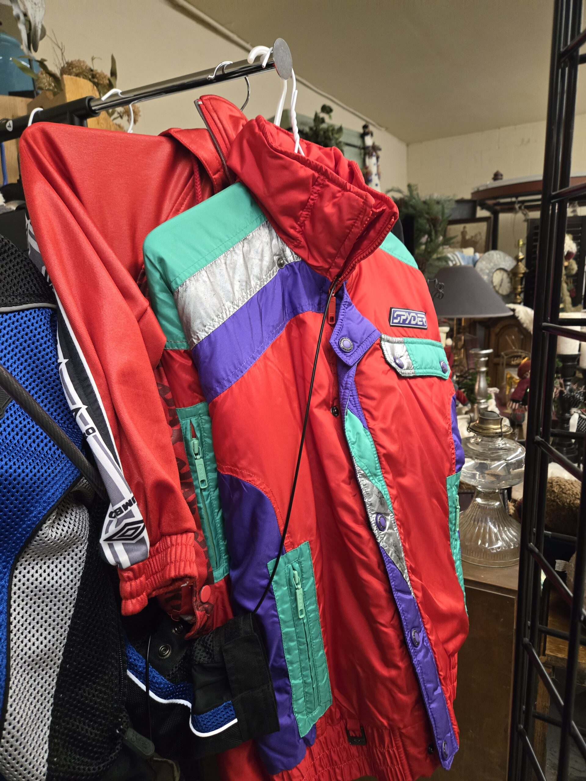 Vintage Spyder ski jacket in red with teal, purple, and metallic silver color blocking, a striped Spyder logo badge, a purple snap placket, a snap-flap chest pocket, and teal zippered pockets, beside a red Umbro track jacket with the Umbro diamond logo on white sleeve taping and a blue, black, and silver Tour Master mesh motorcycle jacket at Shop 89, Pacific Antiques Mall in Tacoma, WA.